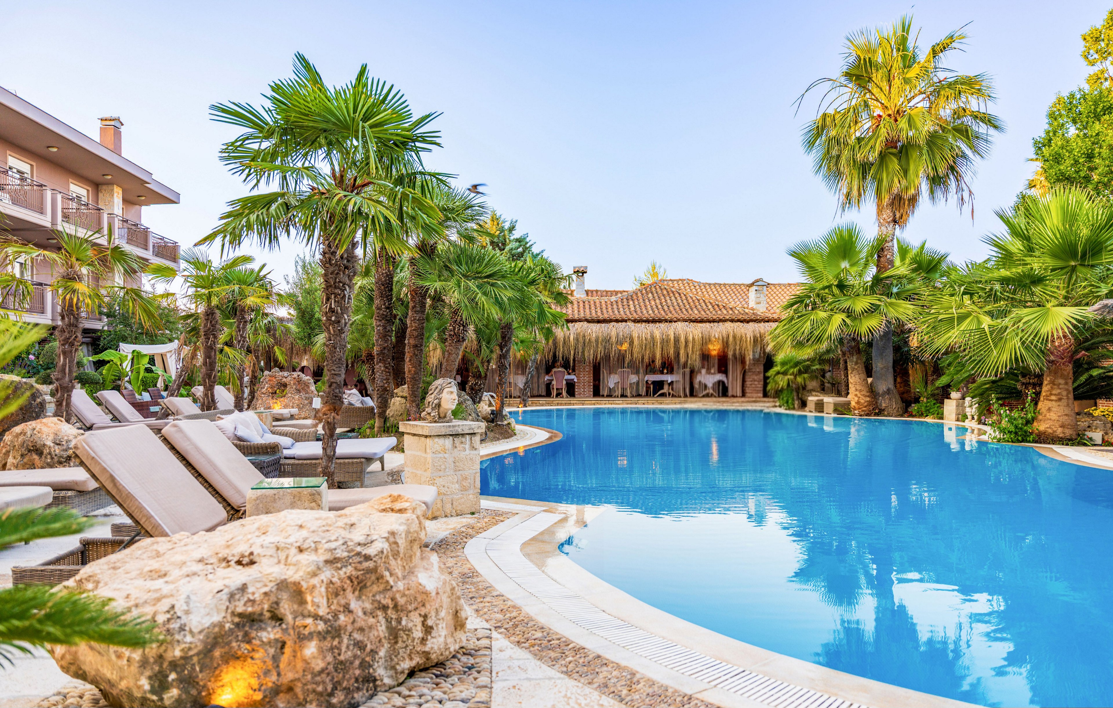 A large curved swimming pool is surrounded by lush palm trees and tropical landscaping at a resort. Sun loungers with cushions line the stone poolside, facing the water for relaxation. In the background, a thatched-roof building and outdoor seating area create a vacation atmosphere. The clear blue sky and reflections in the pool emphasize a warm, tranquil setting.