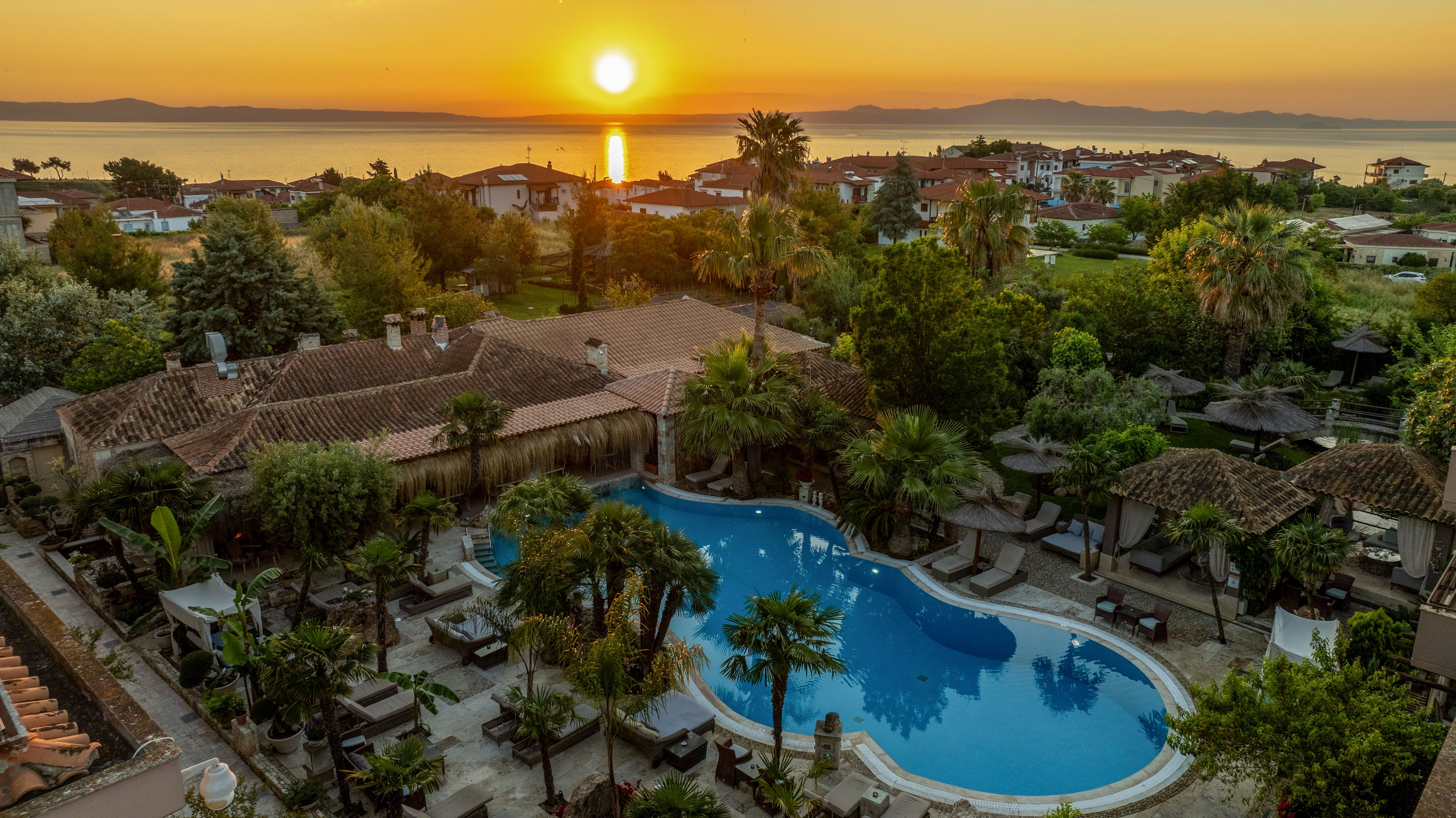 An elevated view shows a resort swimming pool surrounded by lush greenery and tiled-roof buildings. The sun sets over a calm sea in the background, casting warm golden light across the landscape. Palm trees and shaded lounge areas frame the curved pool, creating a tranquil vacation atmosphere. Distant hills and coastal houses complete the scenic seaside setting.