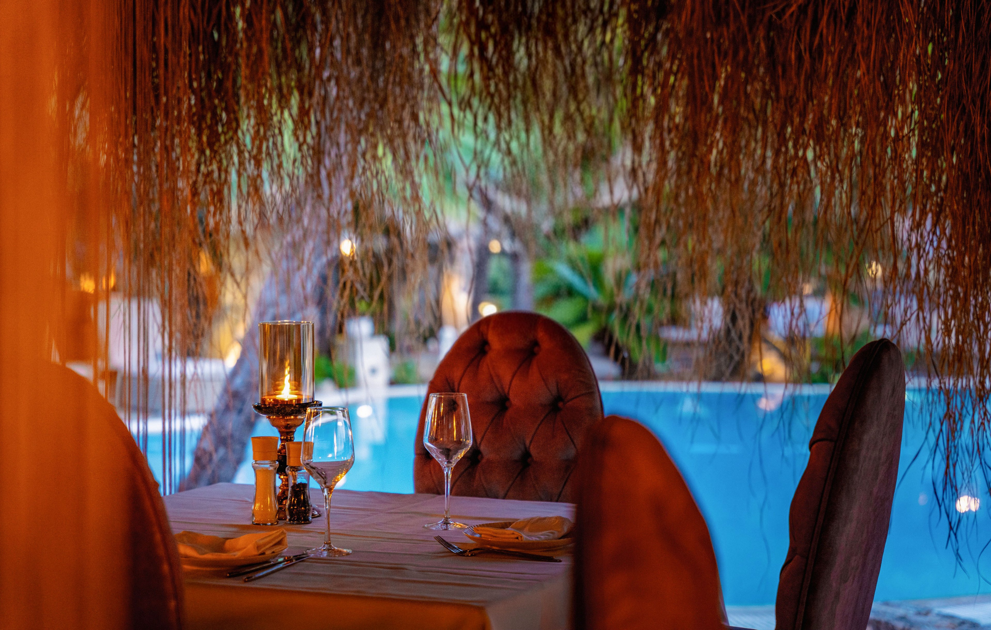 An elegant restaurant table is set for two beside a bright blue swimming pool. Plush upholstered chairs surround a white tablecloth with neatly arranged plates, cutlery, and glassware. A candle lantern adds warm, intimate lighting under a thatched roof, contrasting with the cool tones of the water. Soft evening ambiance and blurred greenery in the background create a tranquil, romantic atmosphere.