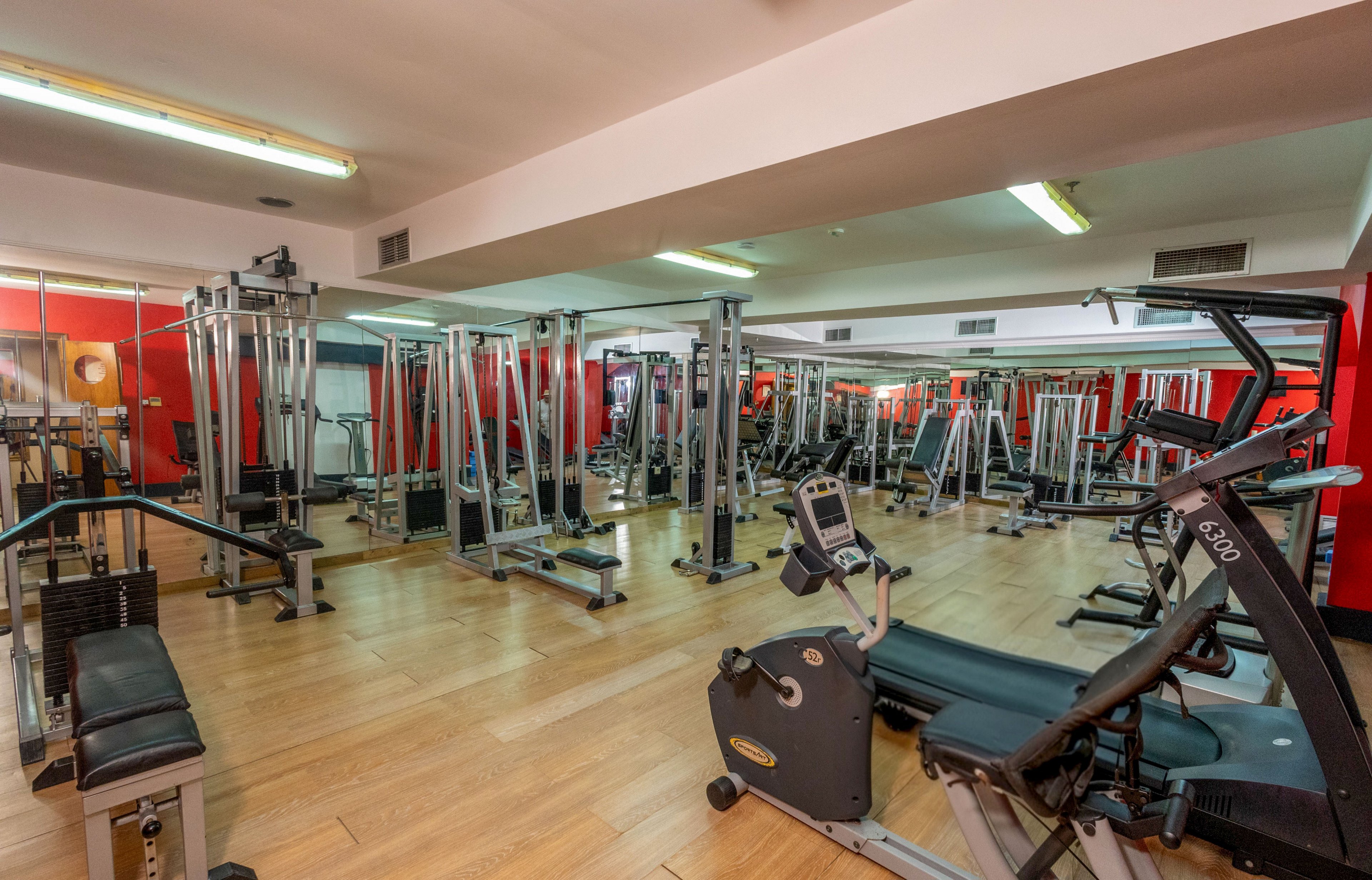 A modern indoor gym is filled with various exercise machines and strength training equipment. The room features wooden flooring, red accent walls, and large mirrors that visually expand the space. Treadmills, weight benches, and cable machines are neatly arranged, creating an organized workout environment. The gym is empty, emphasizing the clean, well-maintained, and ready-for-use fitness area.