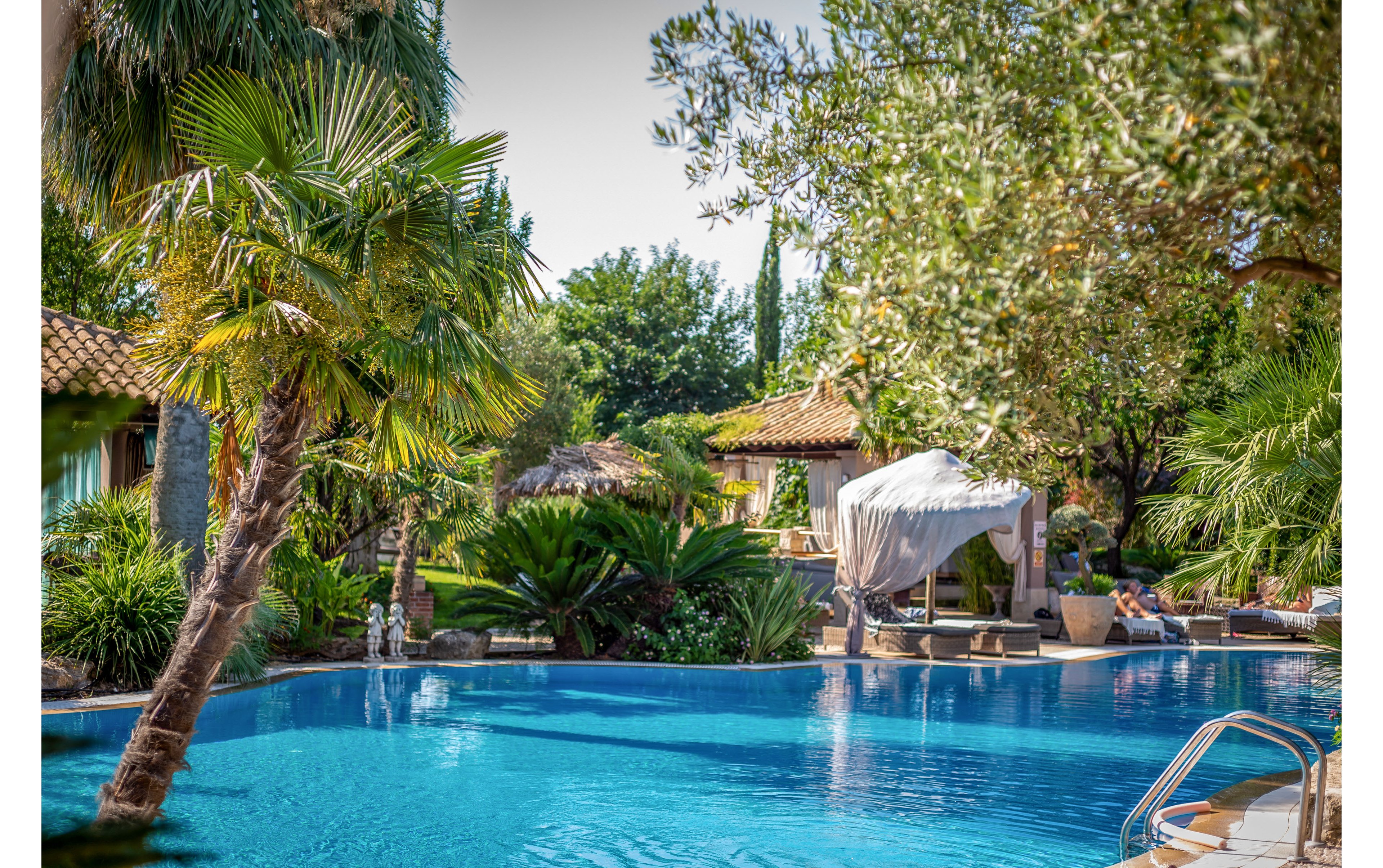 A clear blue swimming pool sits in the foreground of a lush tropical garden. Palm trees and dense greenery surround a rustic villa with tiled roofs, creating a secluded resort atmosphere. Sun loungers and shaded seating areas suggest a relaxing vacation setting. Bright sunlight and reflections on the water enhance the warm, tranquil mood of the scene.