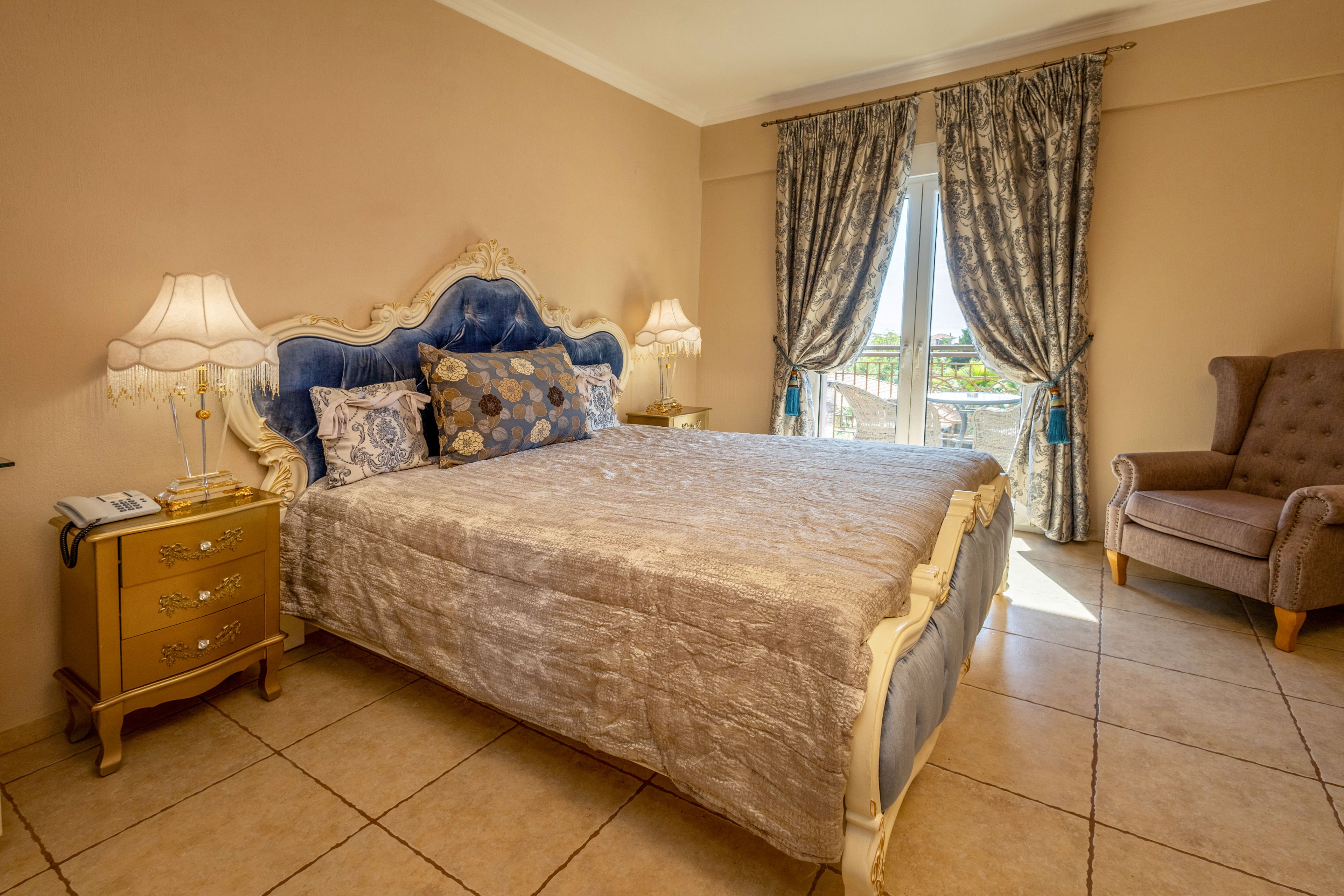 A spacious bedroom features a large upholstered bed with a tufted headboard and decorative pillows. Matching bedside tables with ornate lamps flank the bed, adding a warm glow to the room. Floor-to-ceiling patterned curtains frame a glass door that opens to a bright outdoor view. A cushioned armchair sits in the corner, and the tiled floor and neutral walls create a calm, traditional atmosphere.