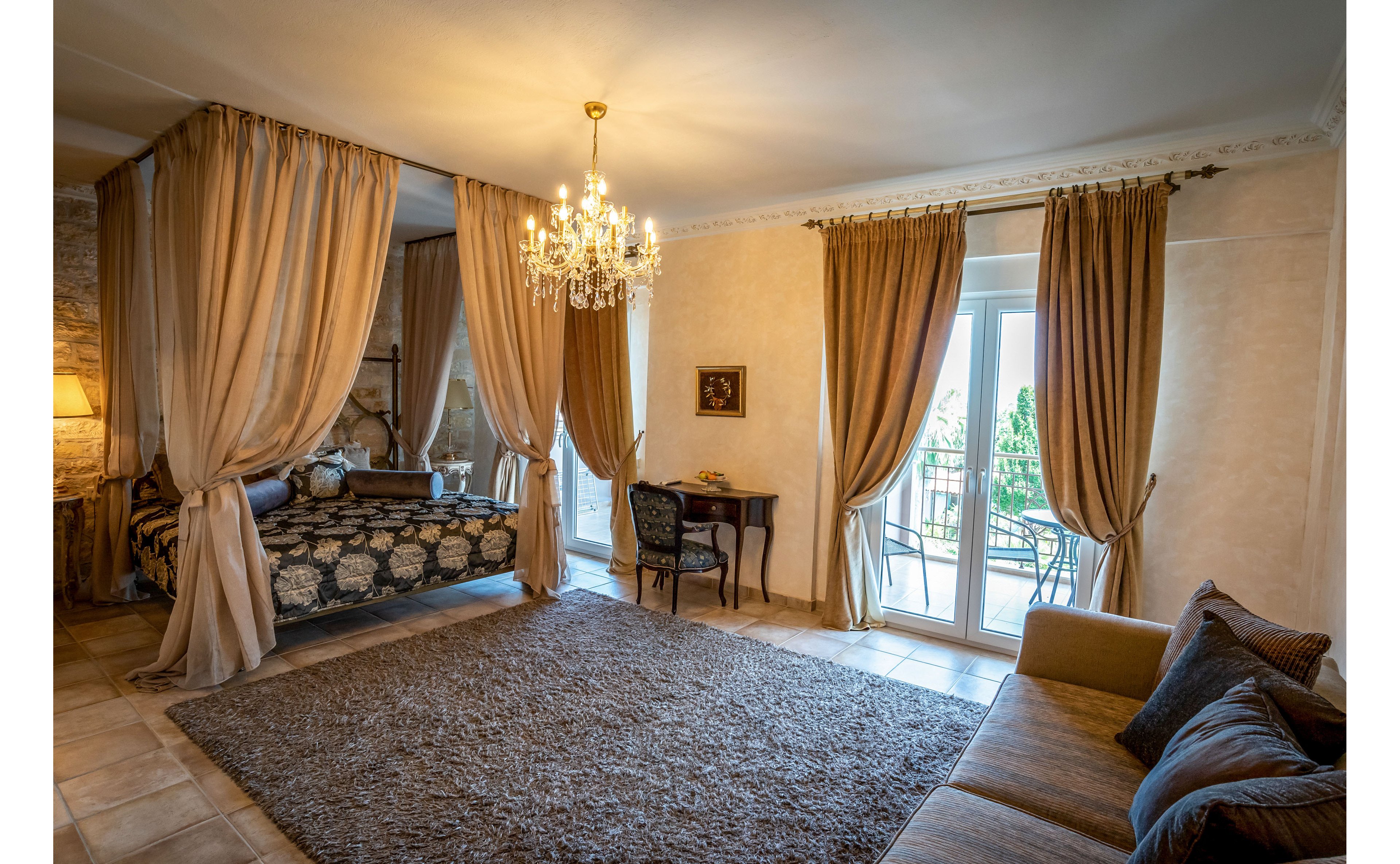 A spacious, elegant bedroom features a canopy bed framed by flowing curtains and a crystal chandelier overhead. Warm beige walls and tiled flooring complement the large textured rug and upholstered sofa. Floor-to-ceiling drapes frame glass doors that open onto a small balcony with outdoor chairs. A compact writing desk and chair sit near the window, adding a functional touch to the luxurious interior.