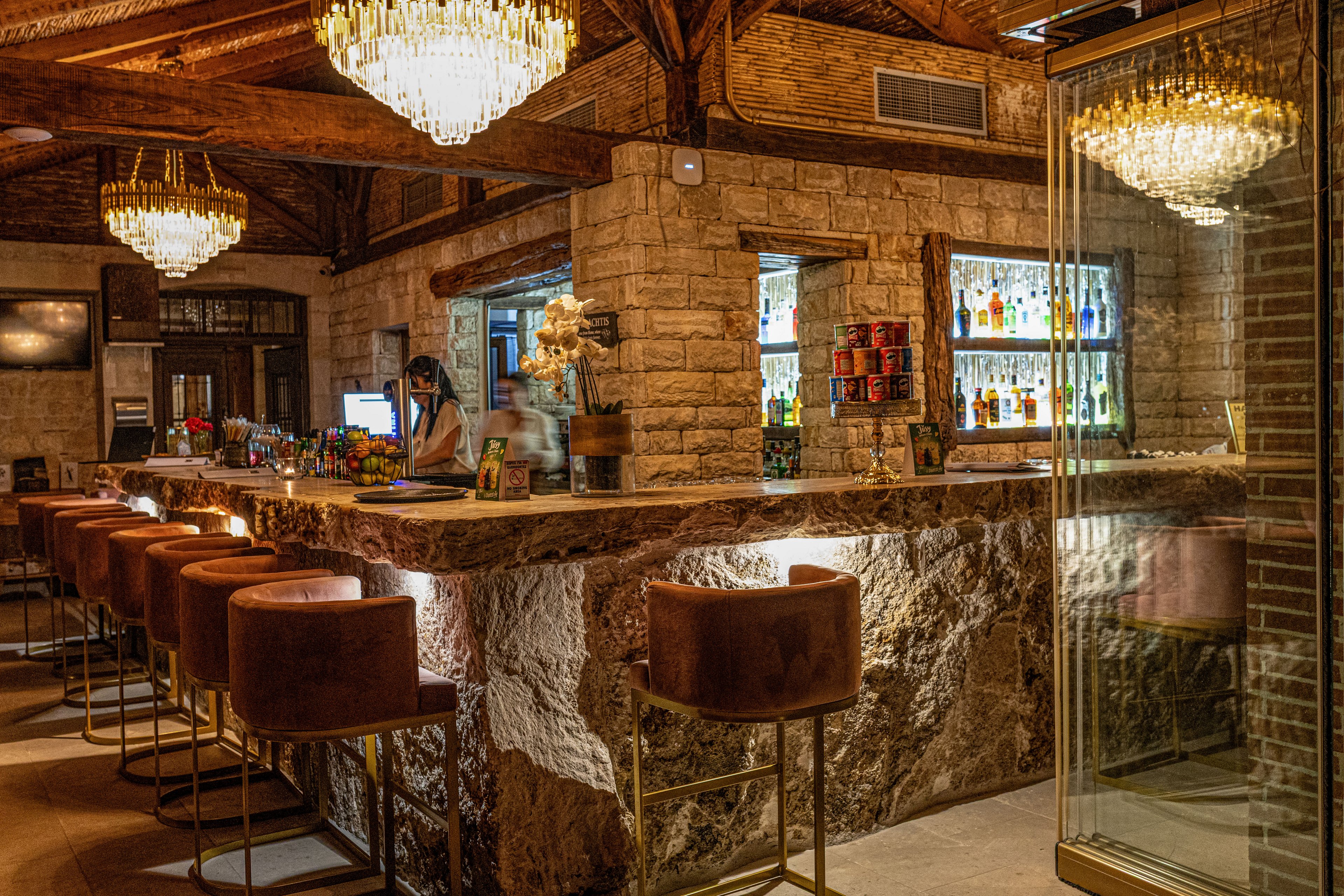 Warmly lit bar interior features a long stone counter with cushioned high stools. Shelves of assorted liquor bottles glow behind the bar, adding color and depth to the rustic stone walls. Elegant crystal chandeliers hang from a wooden beam ceiling, casting a golden light across the space. A bartender station with a computer screen and floral decor completes the inviting, upscale atmosphere.