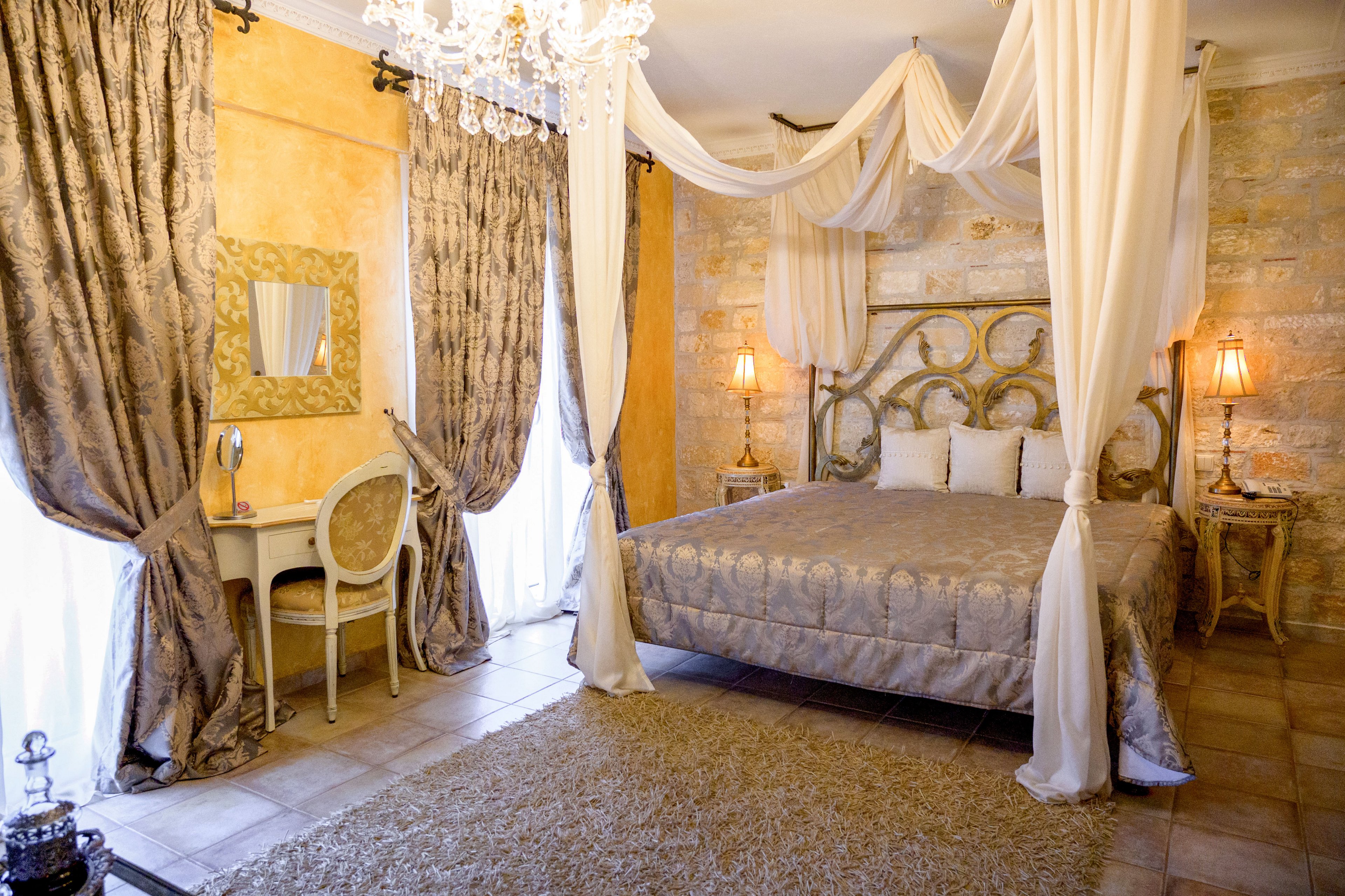 Luxurious bedroom featuring a metal canopy bed draped with sheer white fabric. The room has stone-textured walls, tall patterned curtains, and a soft beige rug on the tiled floor. A small vanity table with a round mirror and chair sits beside the bed, complemented by two warm table lamps on ornate nightstands. The overall ambiance is classic, romantic, and softly illuminated.