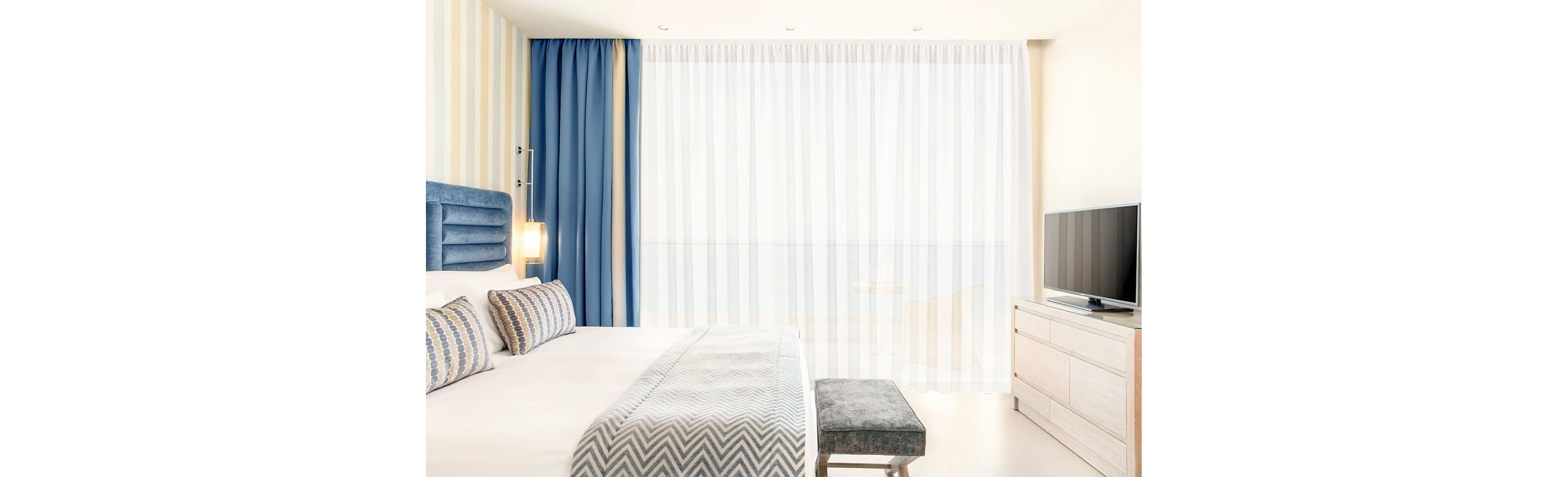 A contemporary hotel bedroom features a neatly made bed with patterned pillows and a matching throw. Floor-to-ceiling sheer curtains cover a large window, allowing soft natural light to fill the room. Blue accents on the headboard and drapes contrast with the neutral walls and furniture. A flat-screen television sits on a light wood dresser beside a small upholstered bench at the foot of the bed.
