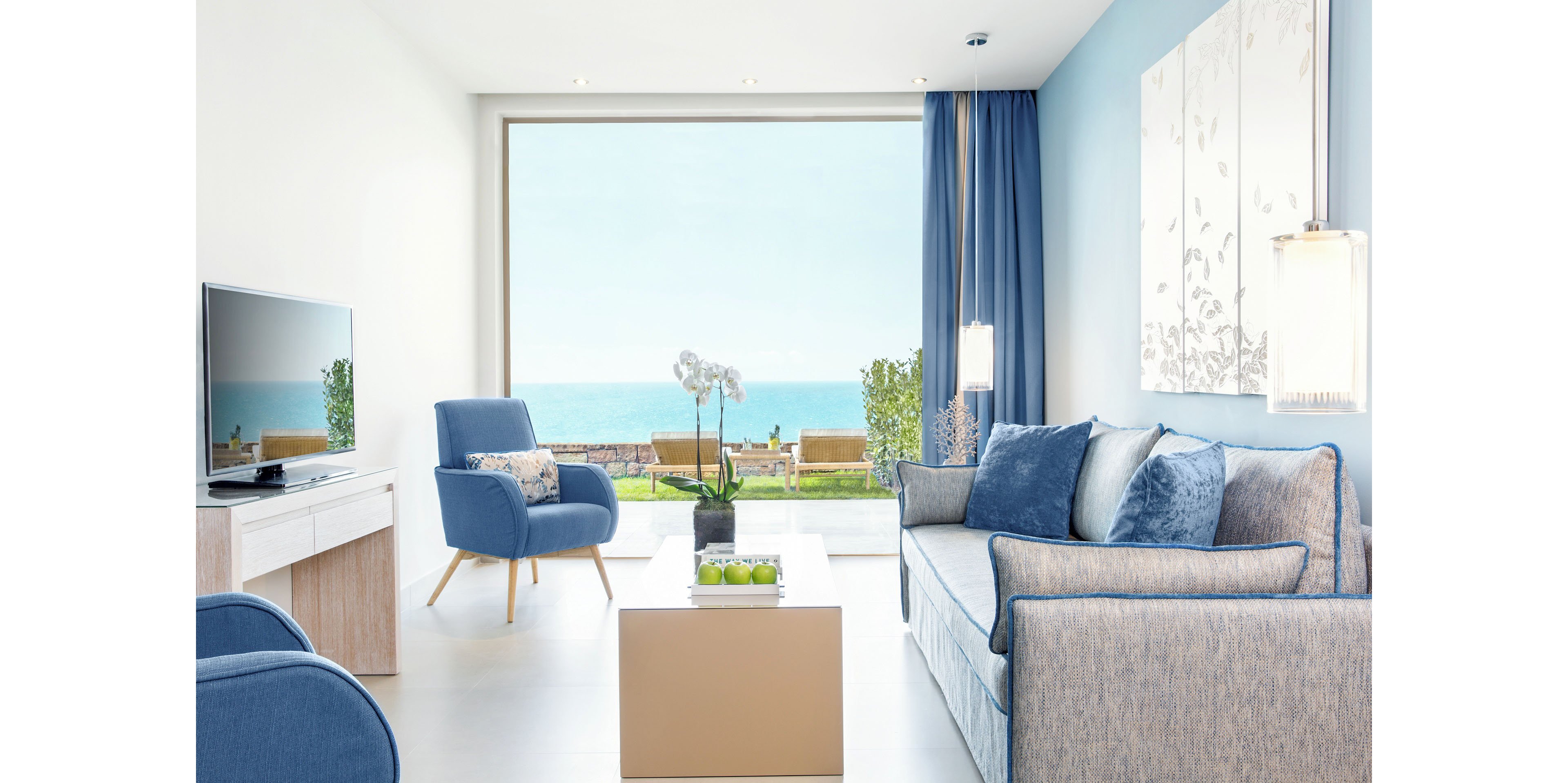A modern living room features a large picture window overlooking a bright blue ocean and outdoor seating area. The interior is decorated in soft blue and white tones, with a sofa, armchair, and minimalist coffee table. A flat-screen television sits on a low console, while sheer curtains and hanging lamps add a light, airy feel. Potted plants and a white orchid bring touches of nature into the contemporary coastal space.