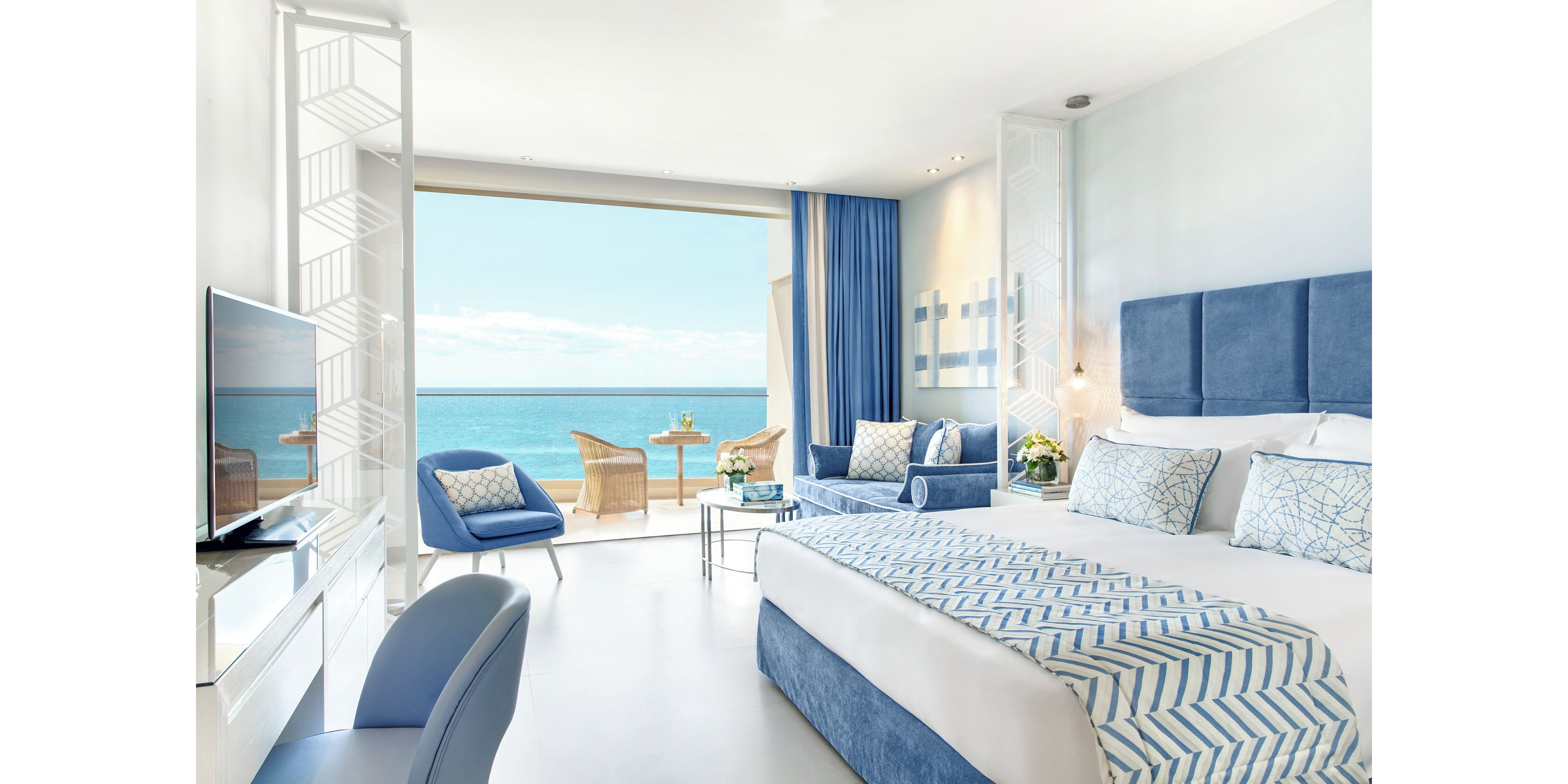 A modern hotel room features a large bed with blue and white geometric bedding and matching upholstered headboard. Floor-to-ceiling glass doors open to a balcony overlooking a calm ocean and clear sky. The interior decor is dominated by soft blue and white tones, with a desk, chair, armchairs, and fresh flowers adding comfort and elegance. Natural light floods the space, creating an airy, relaxing seaside atmosphere.