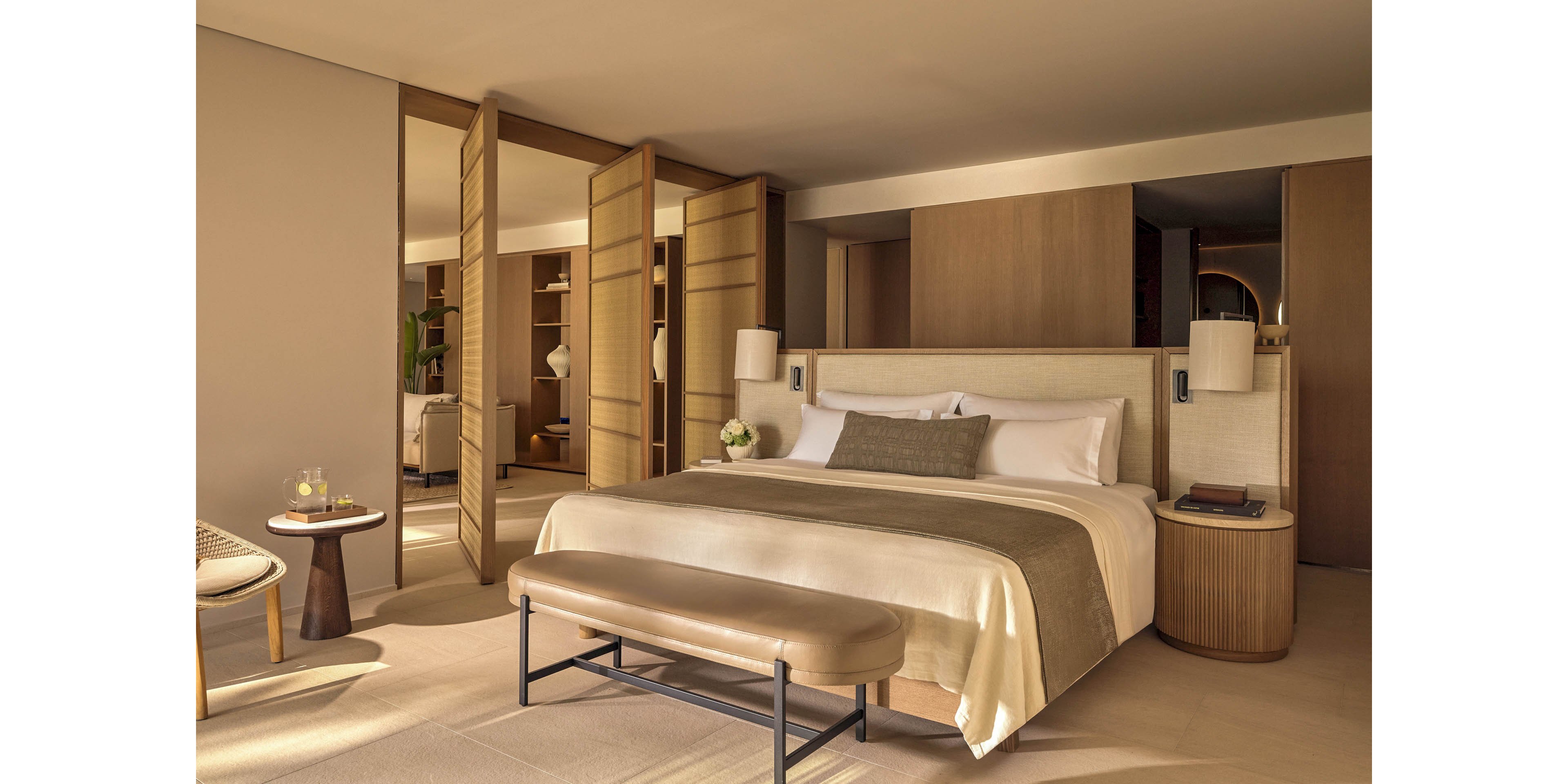 A spacious contemporary bedroom features a large bed with crisp white linens and beige accents. Sliding wooden panels open to reveal an adjoining area, enhancing the suite’s airy, open-plan feel. Warm wood furniture, including a padded bench and bedside tables, complements the soft neutral color palette. Soft lighting and minimal decor create a calm, sophisticated atmosphere ideal for relaxation.
