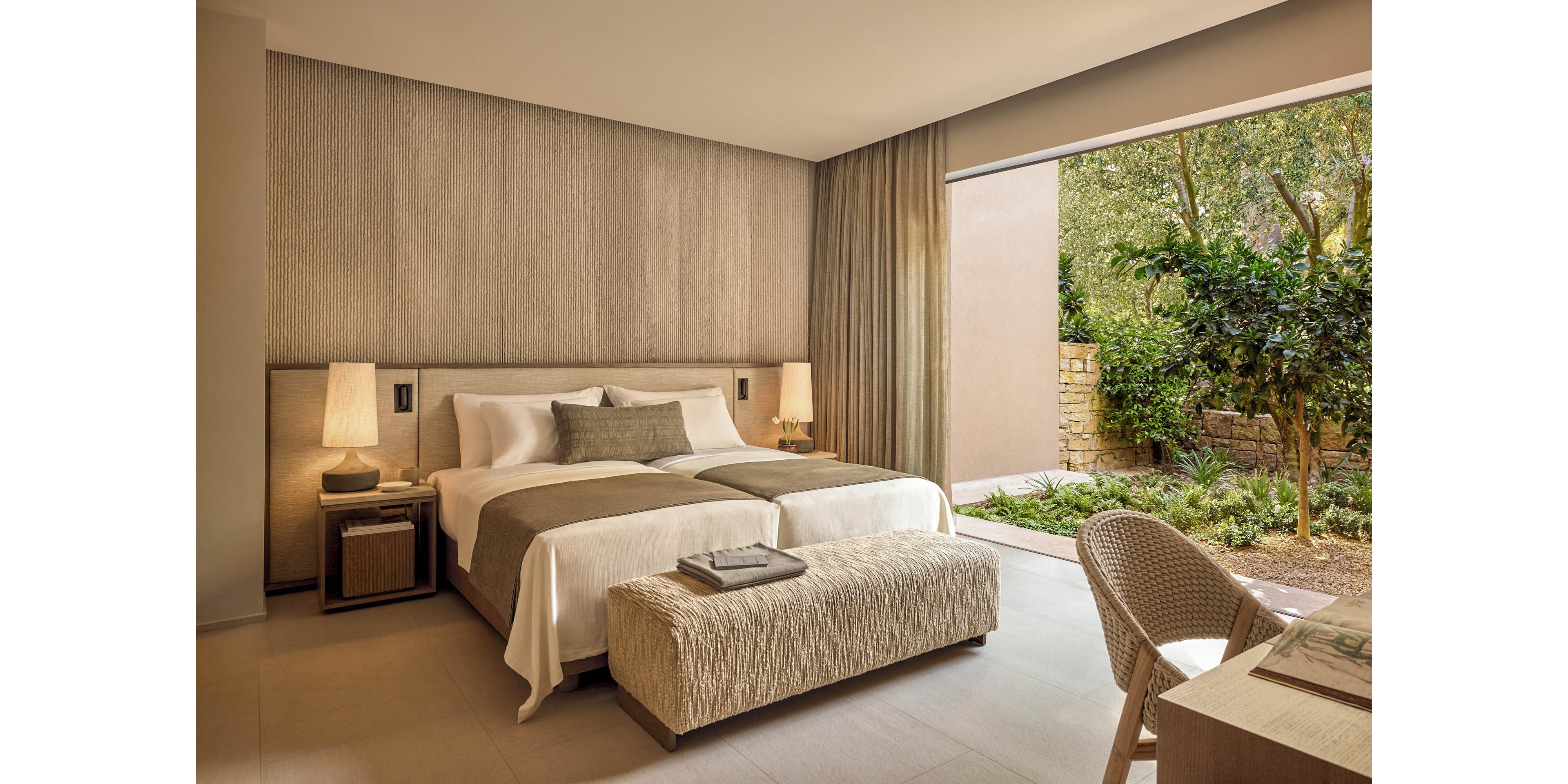 A contemporary hotel bedroom features a large neatly made bed with neutral-toned linens and an upholstered bench at the foot. Warm bedside lamps and textured walls create a cozy, minimalist atmosphere. Floor-to-ceiling curtains frame a wide opening to a lush private garden with trees and greenery. A simple desk and woven chair sit near the window, enhancing the room’s serene, resort-like feel.
