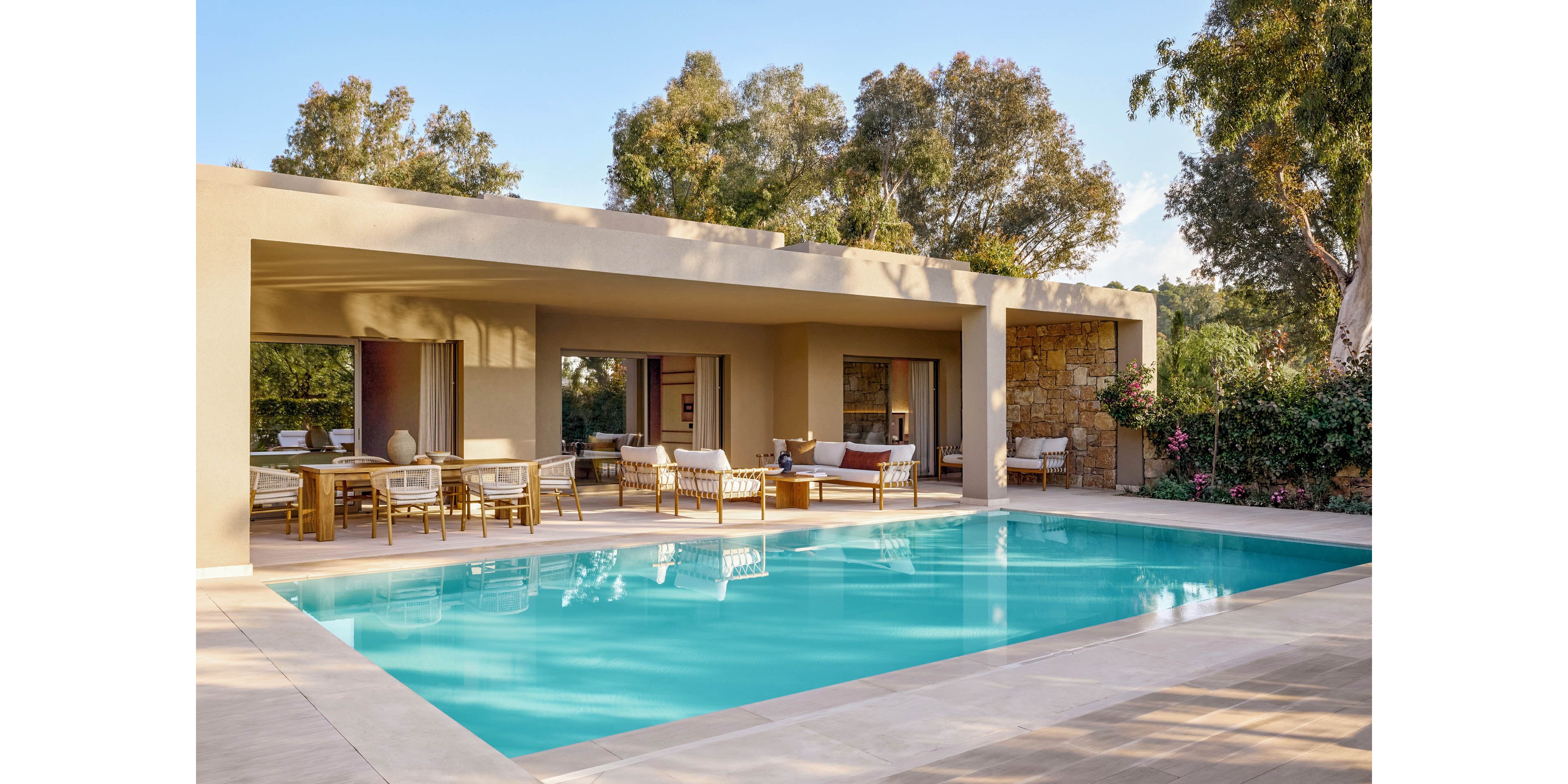 A modern villa features a rectangular outdoor swimming pool surrounded by a light stone terrace. The open-plan patio includes dining and lounge areas furnished with neutral-toned chairs and sofas. Large openings connect the indoor and outdoor spaces, emphasizing a relaxed, resort-style atmosphere. Lush trees and greenery frame the scene, creating a tranquil and private retreat.