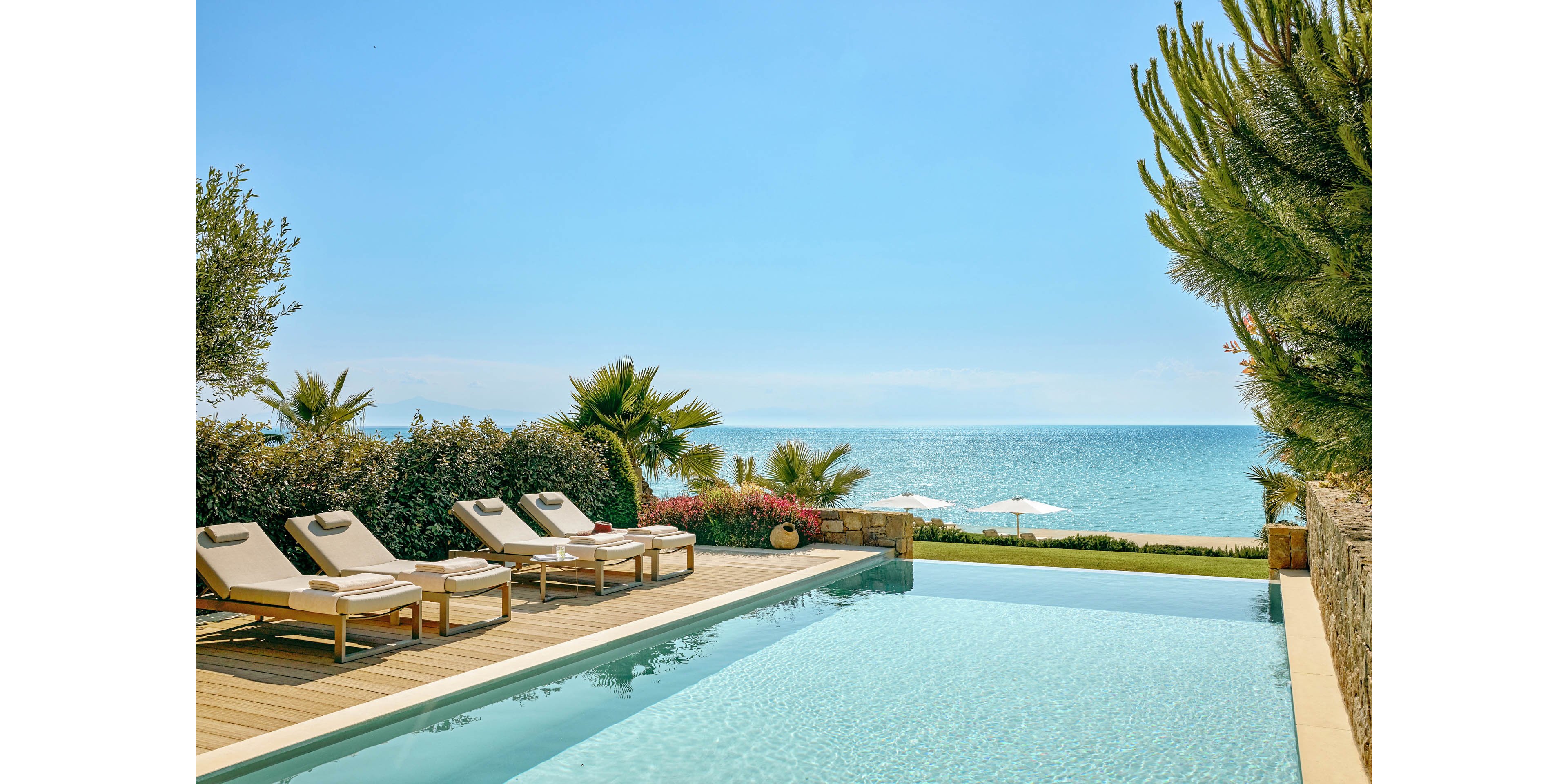 A serene infinity pool overlooks a calm, turquoise sea under a clear blue sky. Several cushioned sun loungers with umbrellas are arranged on a wooden deck beside the pool. Lush greenery, including palm trees and shrubs, frames the scene and adds a tropical feel. The bright sunlight and soft colors create a relaxing, upscale resort atmosphere.