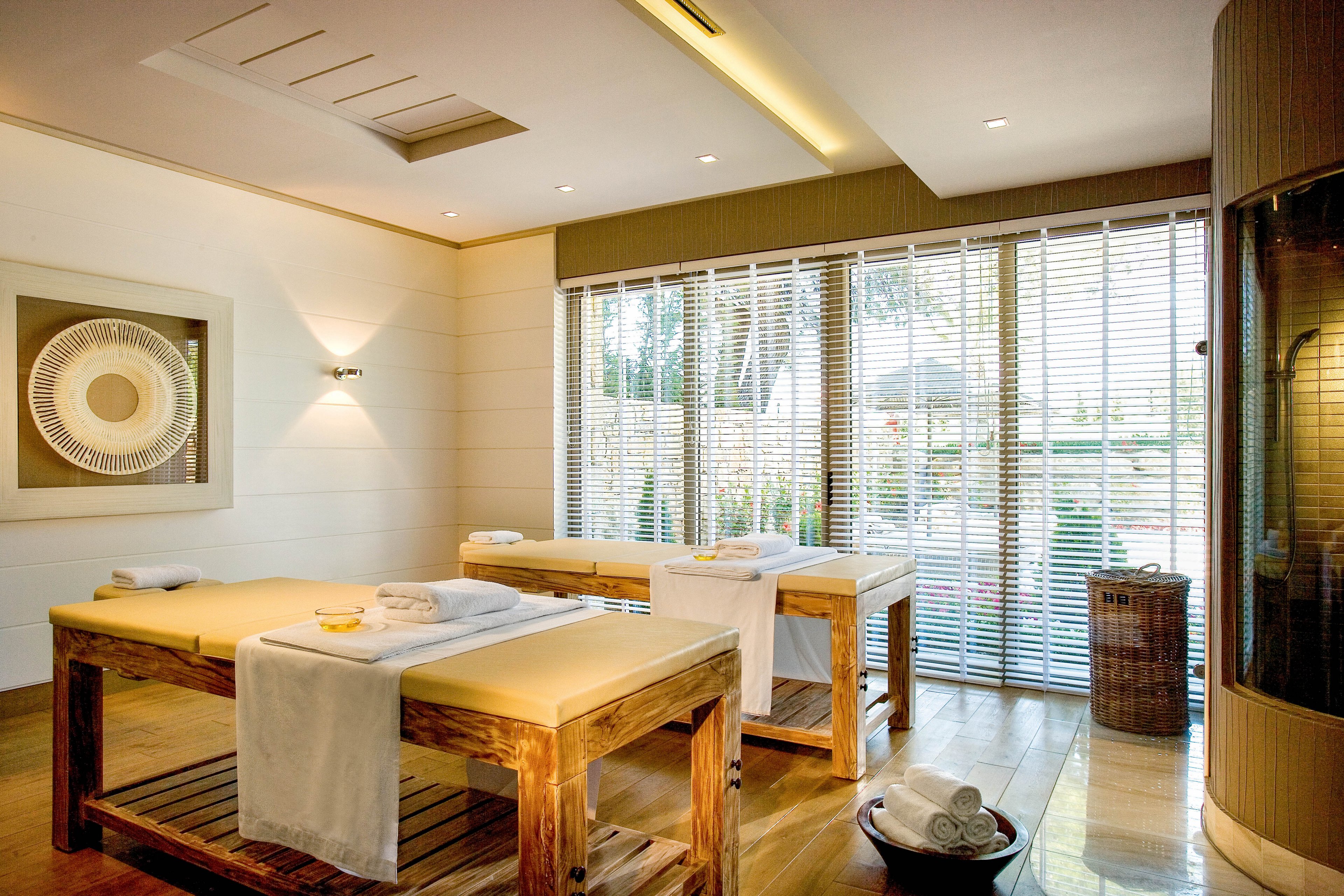 A serene spa treatment room features two wooden massage tables neatly prepared with white towels. Large windows with horizontal blinds let in abundant natural light and offer a view of greenery outside. Warm wooden flooring, neutral walls, and soft lighting create a calm, relaxing atmosphere. Minimal decor, including wall art and spa accessories, enhances the clean and luxurious feel of the space.