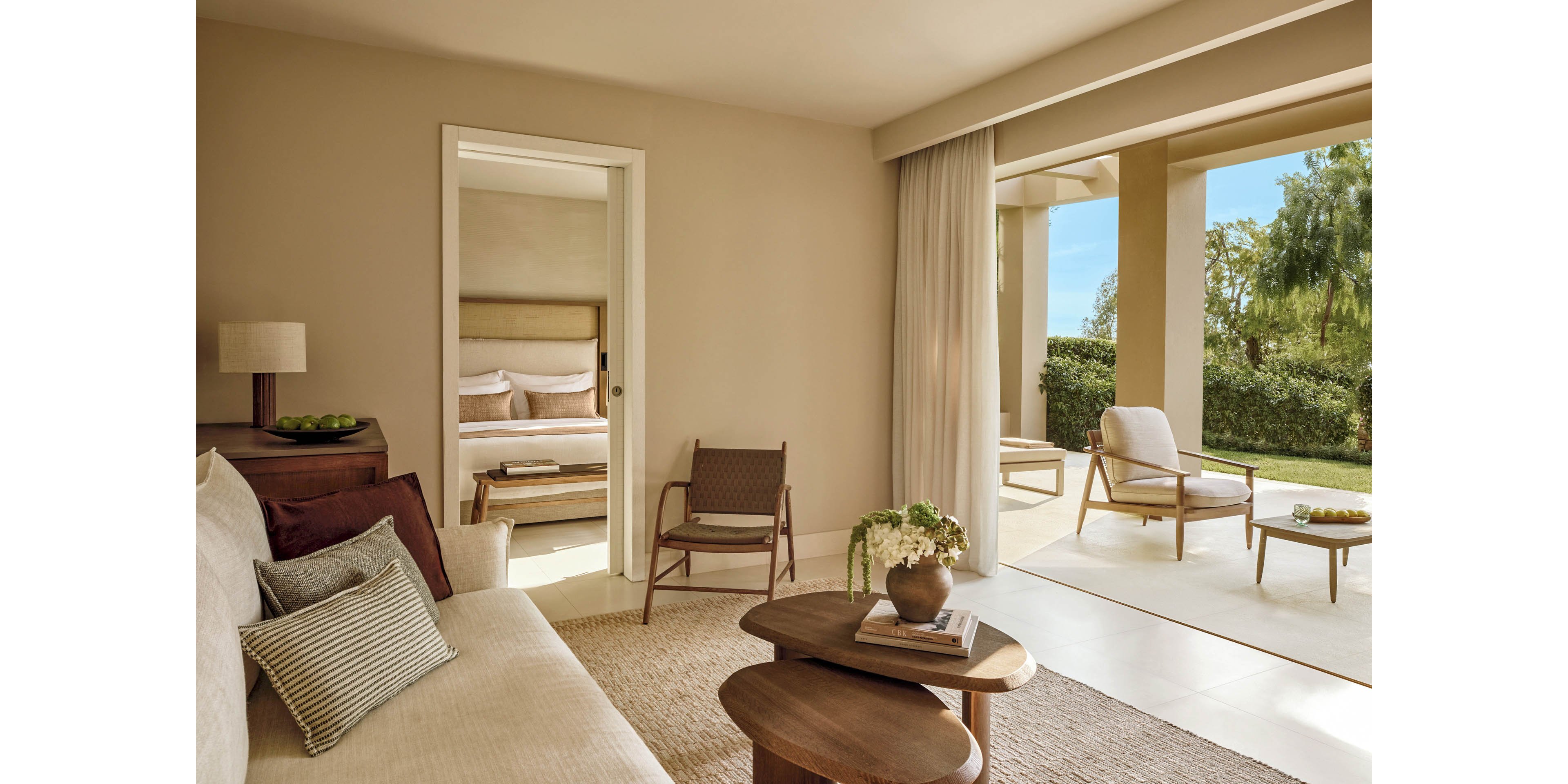A contemporary suite features a cozy living area with a sofa, nested wooden coffee tables, and a woven rug in soft neutral tones. Through an open doorway, a neatly made bed is visible in the adjoining bedroom. Large sliding doors open to a covered terrace with lounge chairs and a view of greenery outside. Natural light and minimalist decor create a calm, luxurious atmosphere ideal for relaxation.