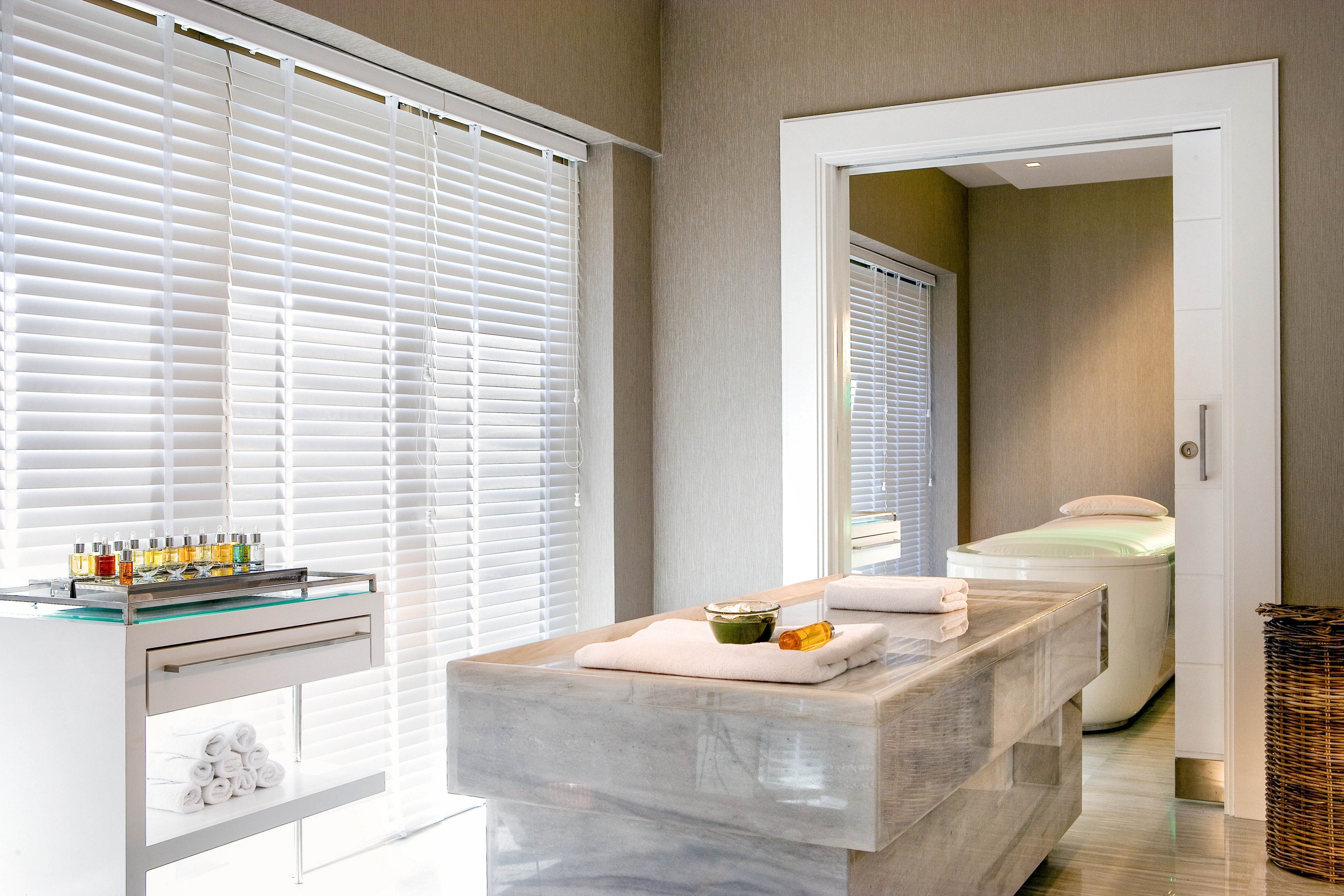 A contemporary spa treatment room features a large stone massage table in the foreground. A second padded treatment bed is visible through an open doorway in an adjoining room. Natural light filters through white window blinds, creating a calm and bright atmosphere. A small trolley with oils and towels and a wicker basket add to the wellness setting.