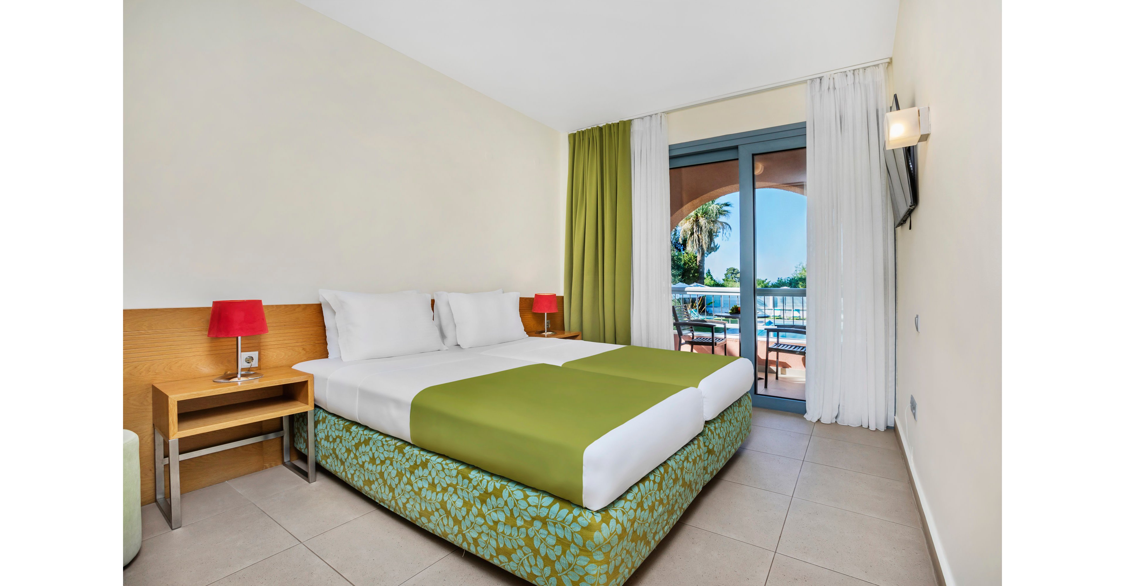 A modern hotel room features two twin beds with white linens and green runners, set against a light-colored wall and tiled floor. A small wooden desk with a red lamp and power outlet sits beside the bed, while a flat-screen TV is mounted on the opposite wall. Floor-to-ceiling curtains frame glass doors that open onto a balcony overlooking a swimming pool and palm trees. The space feels clean, airy, and inviting, with simple contemporary decor and natural light streaming in.