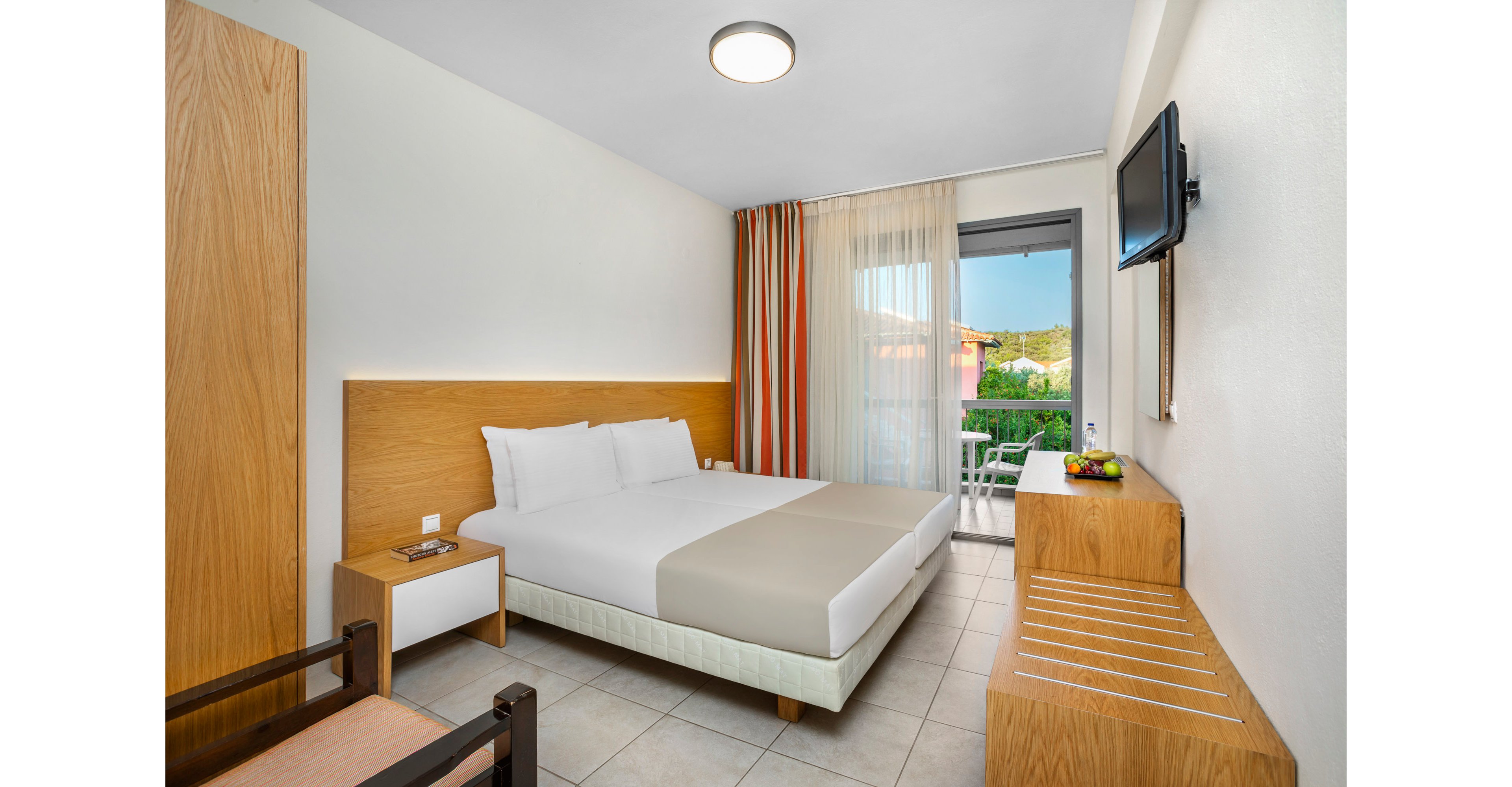 A contemporary hotel room features a double bed with white linens and a light beige runner against a wooden headboard. The space includes a wall-mounted flat-screen TV, a wooden desk with a bowl of fruit, and a bench with slatted top. Floor-to-ceiling curtains in neutral and striped colors frame a glass door leading to a balcony with outdoor scenery. Clean lines, light tones, and minimal decor create a fresh, inviting atmosphere.