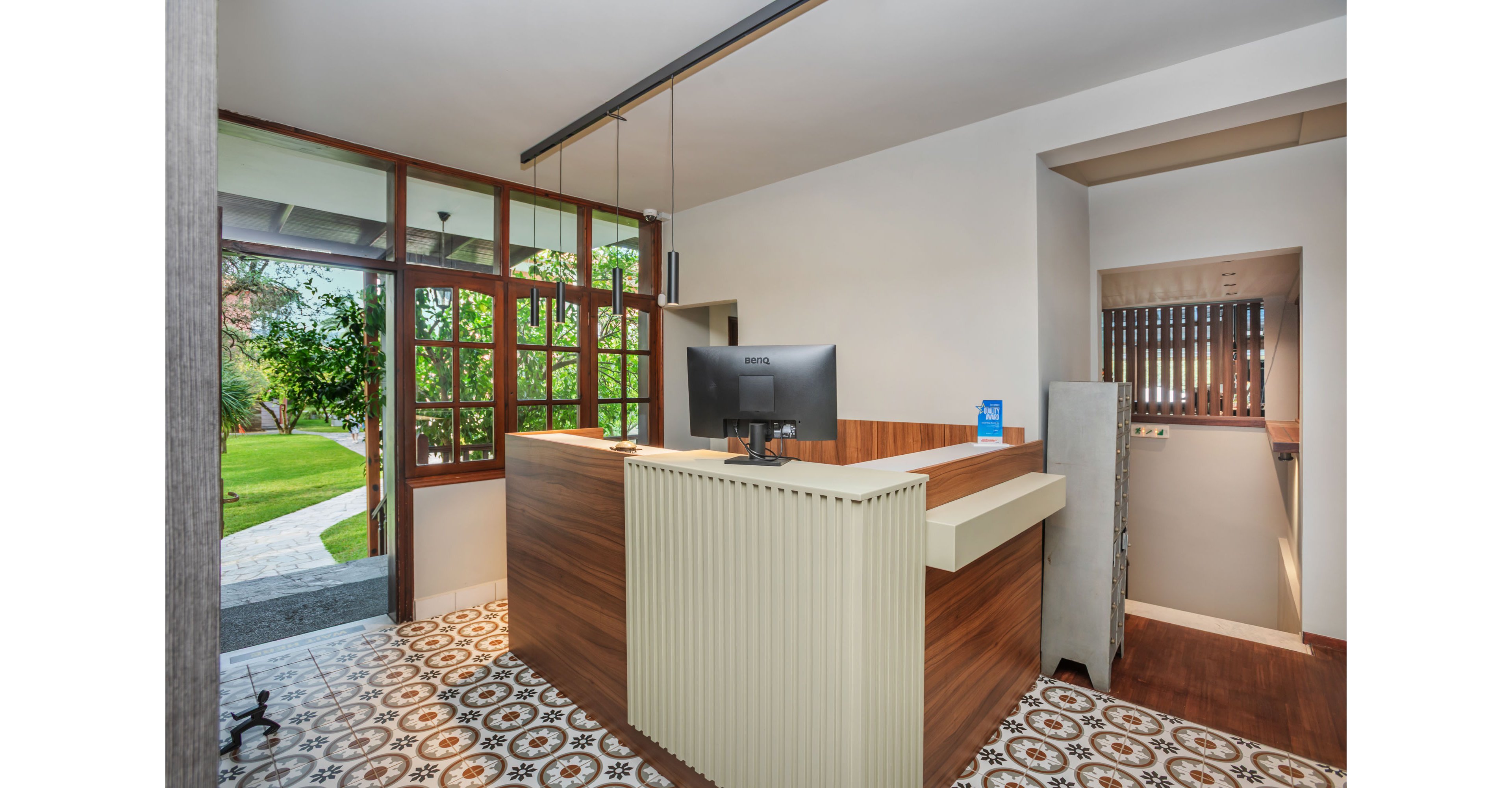 A contemporary reception desk with a computer monitor stands in a bright lobby area. Large glass windows and doors open to a lush green garden outside, allowing natural light to fill the space. The interior features patterned floor tiles, wood paneling, and clean white walls for a modern yet warm look. A small staircase and corridor are visible to the right, adding depth to the architectural layout.