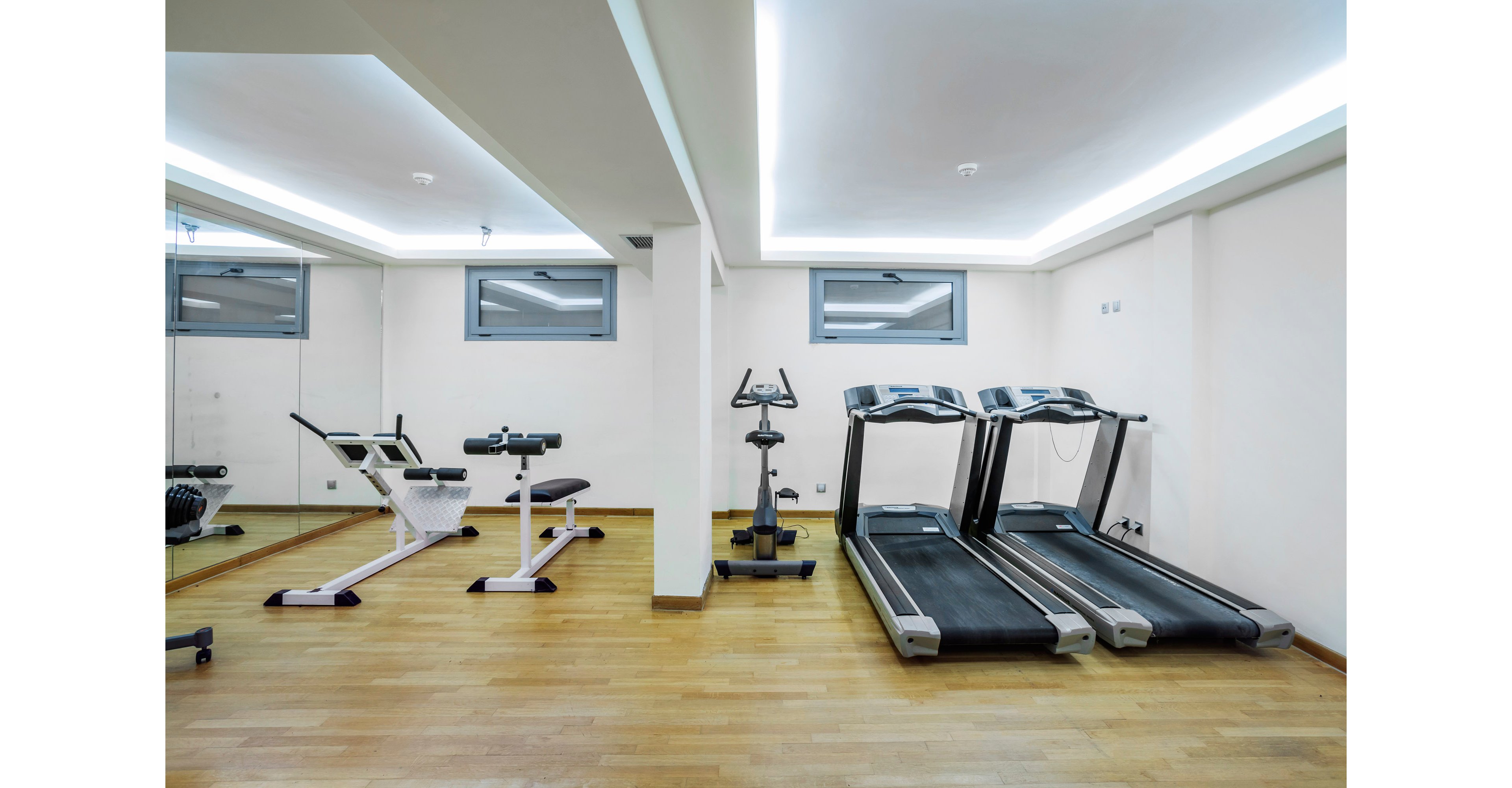 A bright, modern fitness room features two treadmills alongside various exercise machines on a wooden floor. The space is clean and minimal, with white walls and recessed ceiling lighting creating an airy atmosphere. Large mirrors and small horizontal windows add depth and natural light to the room. The overall setting suggests a quiet, well-equipped home or apartment gym ready for cardio and strength workouts.