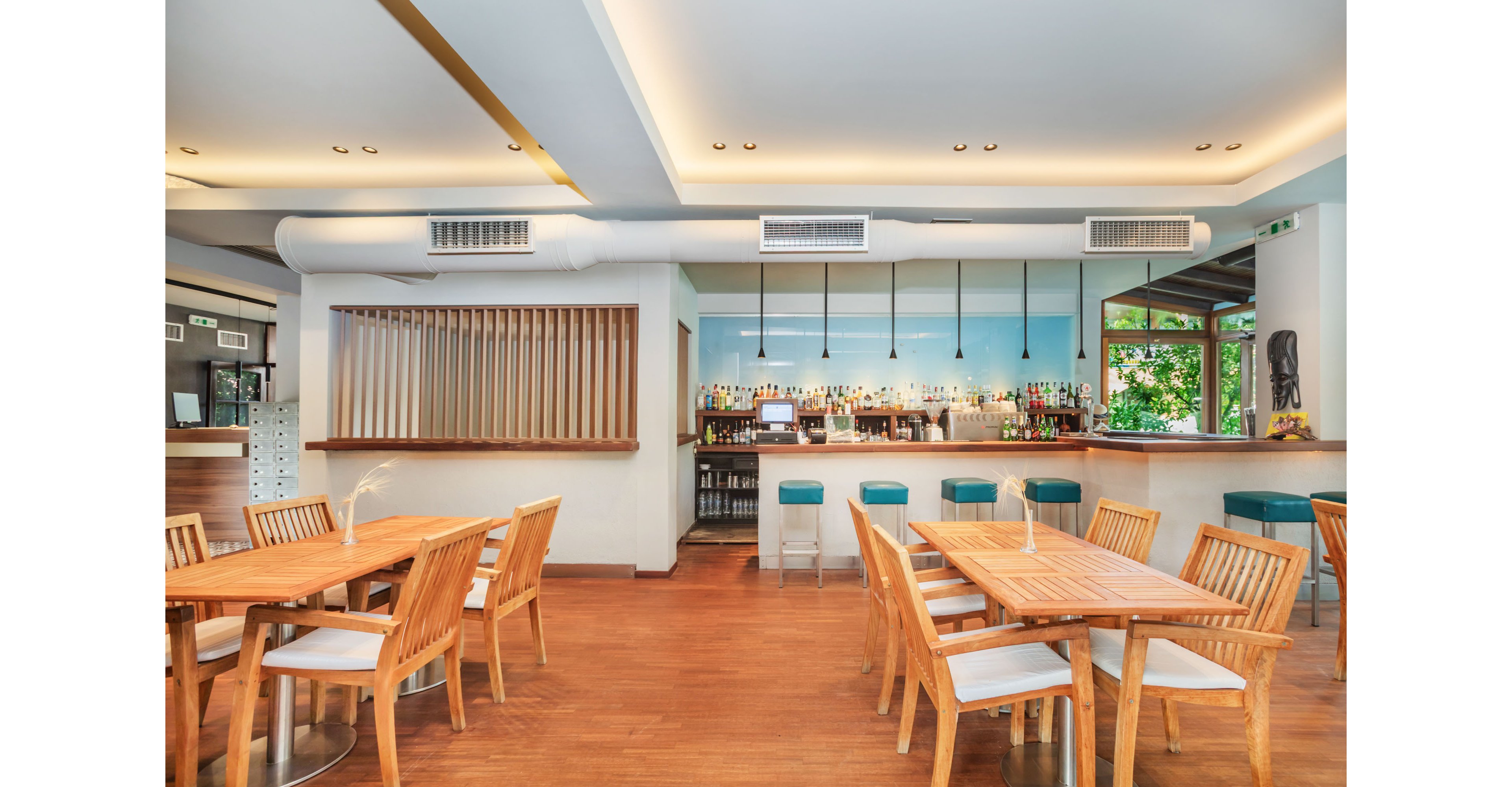 A contemporary restaurant interior features wooden tables and chairs arranged neatly on a warm-toned floor. In the background, a well-stocked bar with high stools and hanging pendant lights creates a focal point. Large windows and glass doors bring in natural light and views of greenery outside. The overall design is clean and minimal, with soft ceiling lighting and simple decorative elements for a relaxed dining atmosphere.