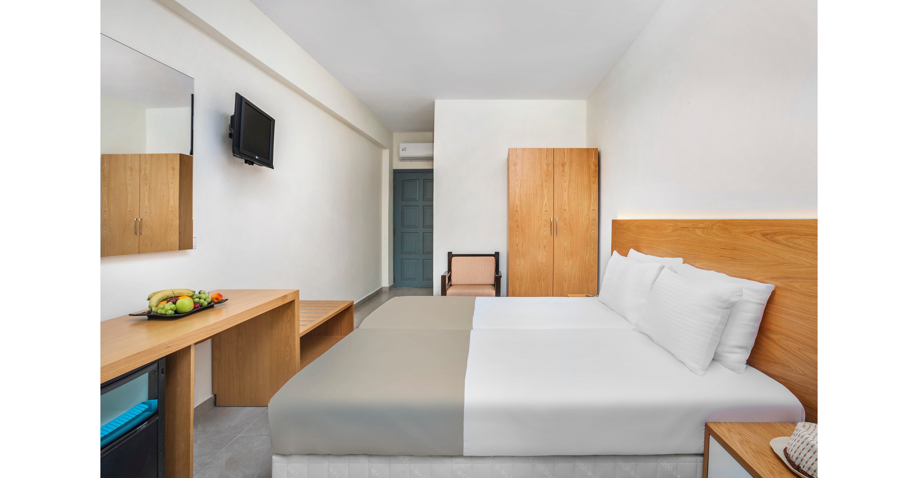 Bright, modern hotel room featuring two neatly made twin beds with white linens and a light gray runner. The space includes a wooden wardrobe, desk, and wall-mounted television, creating a clean and functional layout. A bowl of fresh fruit adds color on the desk, while neutral walls and simple furniture emphasize a minimalist style. The room appears tidy and inviting, suitable for travelers seeking a comfortable stay.