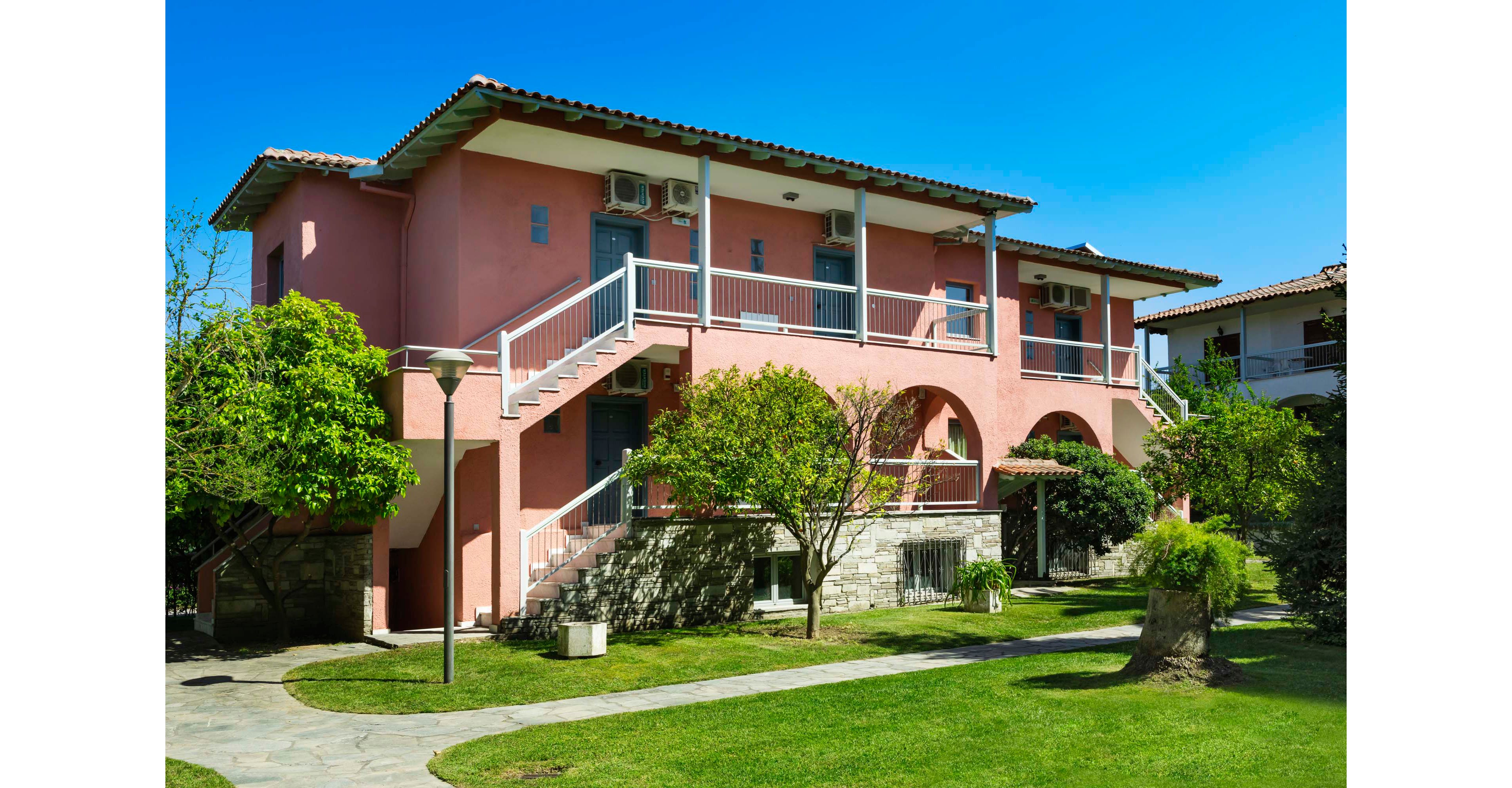 A pink two-story residential building stands in a neatly maintained courtyard. Exterior staircases lead to upper-level balconies with railings and visible air conditioning units. Green trees, trimmed grass, and stone pathways surround the house, creating a peaceful outdoor setting. The clear blue sky and bright daylight emphasize the warm, inviting atmosphere of the property.