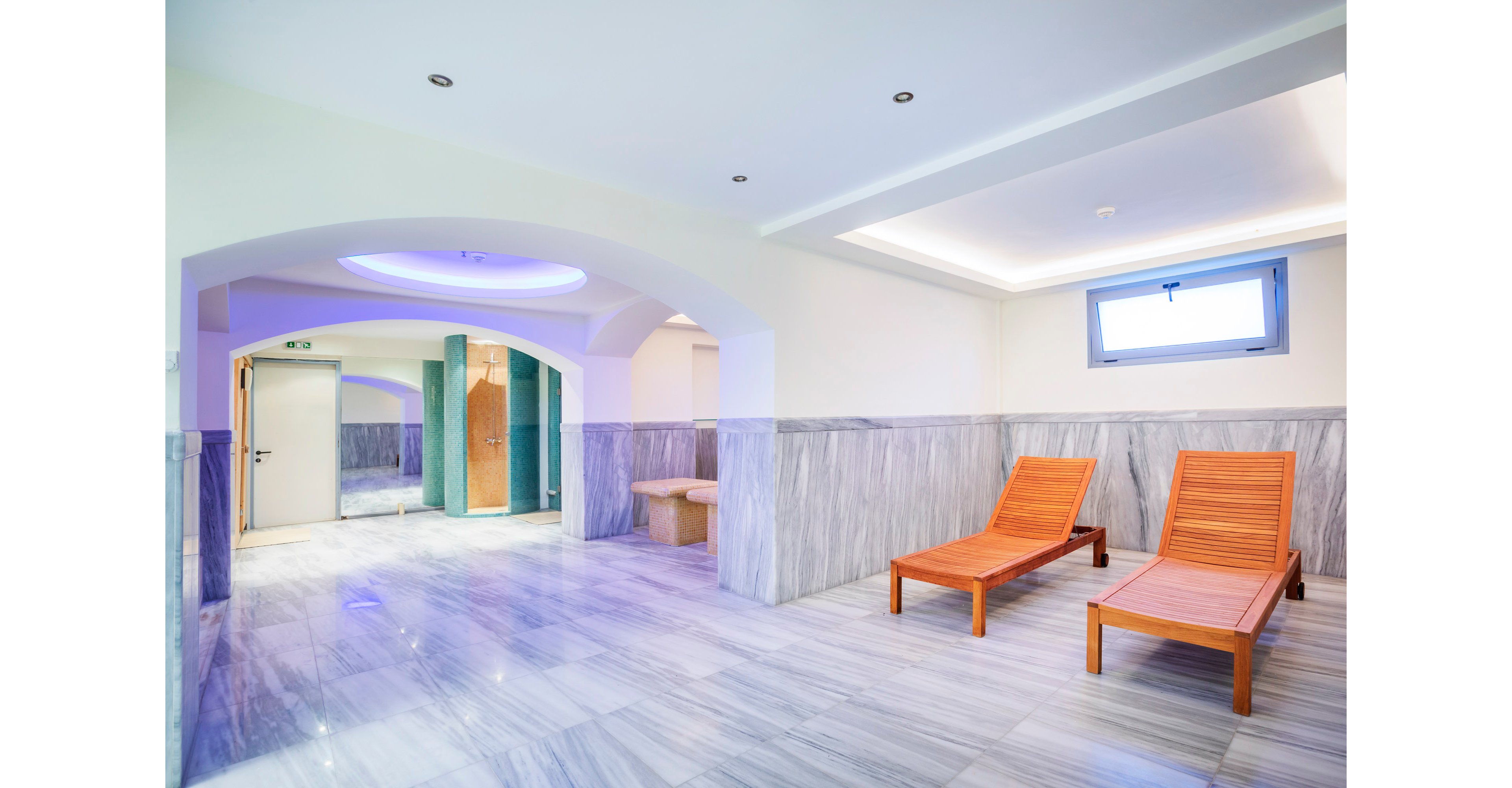A modern spa relaxation room features two wooden lounge chairs on a polished marble floor. The space is bright and minimal, with light-colored walls and a small high window letting in natural light. In the background, an arched opening leads to a shower or steam area with tiled surfaces and soft ceiling lighting. The overall atmosphere is clean, tranquil, and designed for rest and wellness.