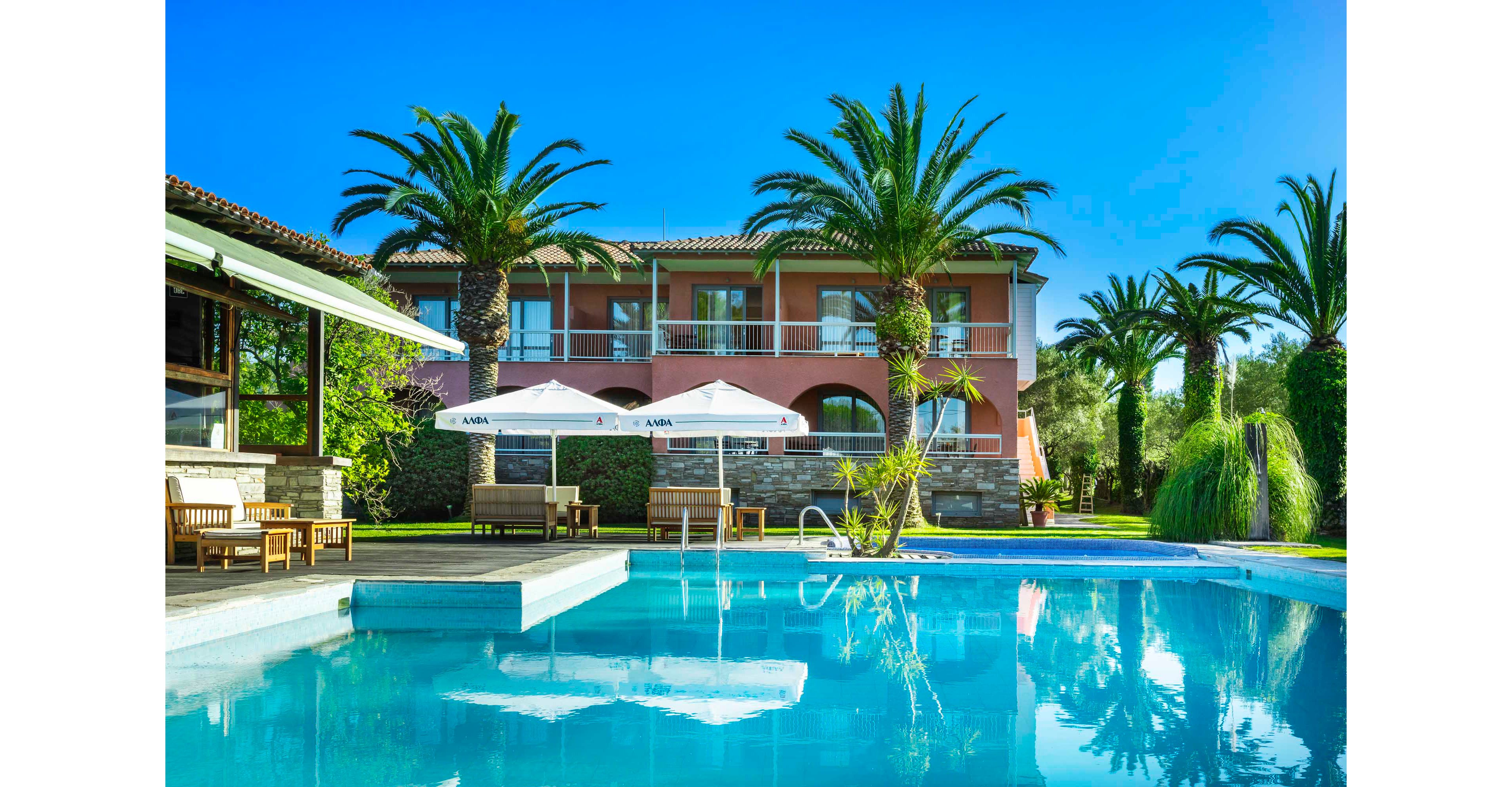A luxurious villa with a large outdoor swimming pool is set amid tall palm trees and lush greenery. Sun loungers and wooden seating areas line the poolside deck, shaded by white umbrellas. The building features warm pastel walls, arched windows, and balconies overlooking the water. Bright blue sky and clear reflections in the pool create a vibrant, relaxing holiday atmosphere.