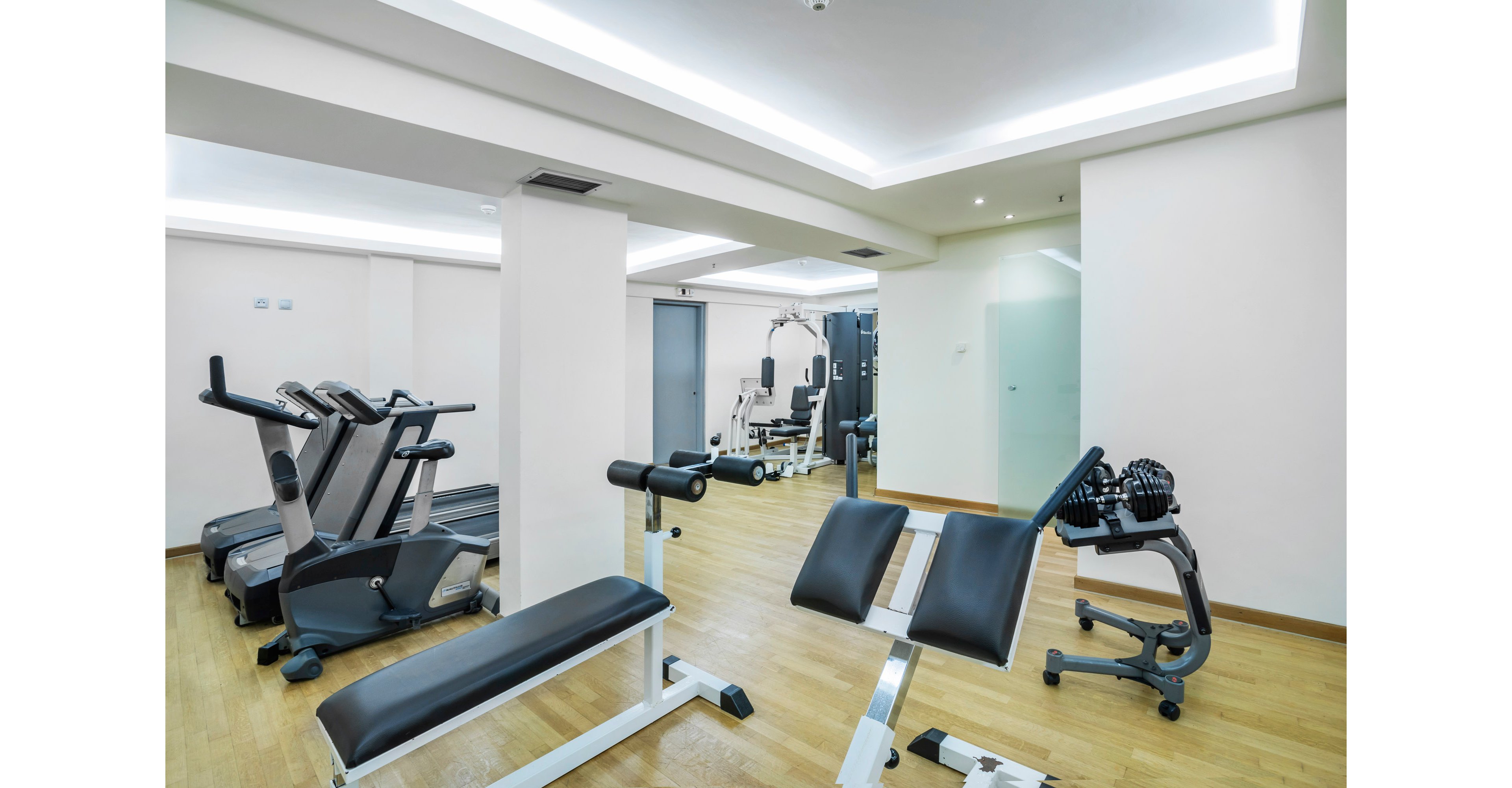 A clean, modern gym interior features various exercise machines and benches arranged on a light wooden floor. Treadmills and strength-training equipment are positioned along the walls, with a central white column dividing the space. Bright, recessed ceiling lighting and white walls create an open, minimalist atmosphere. The room appears empty, emphasizing the equipment and spacious layout.
