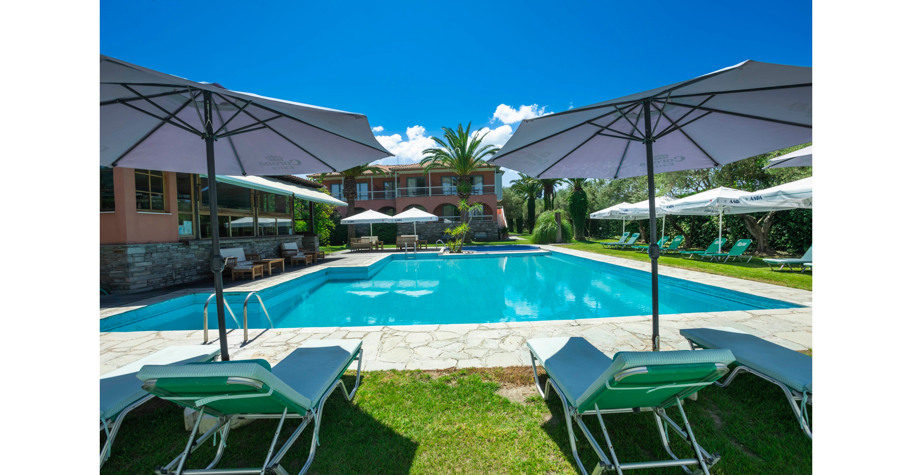 An outdoor swimming pool is surrounded by sun loungers and large white umbrellas in front of a resort-style building. The scene is set on a bright sunny day with a vivid blue sky and scattered white clouds. Green grass and landscaped trees, including a tall palm, frame the pool area, creating a relaxing vacation atmosphere. The clear water and ample seating suggest a comfortable space for guests to unwind and enjoy the warm weather.