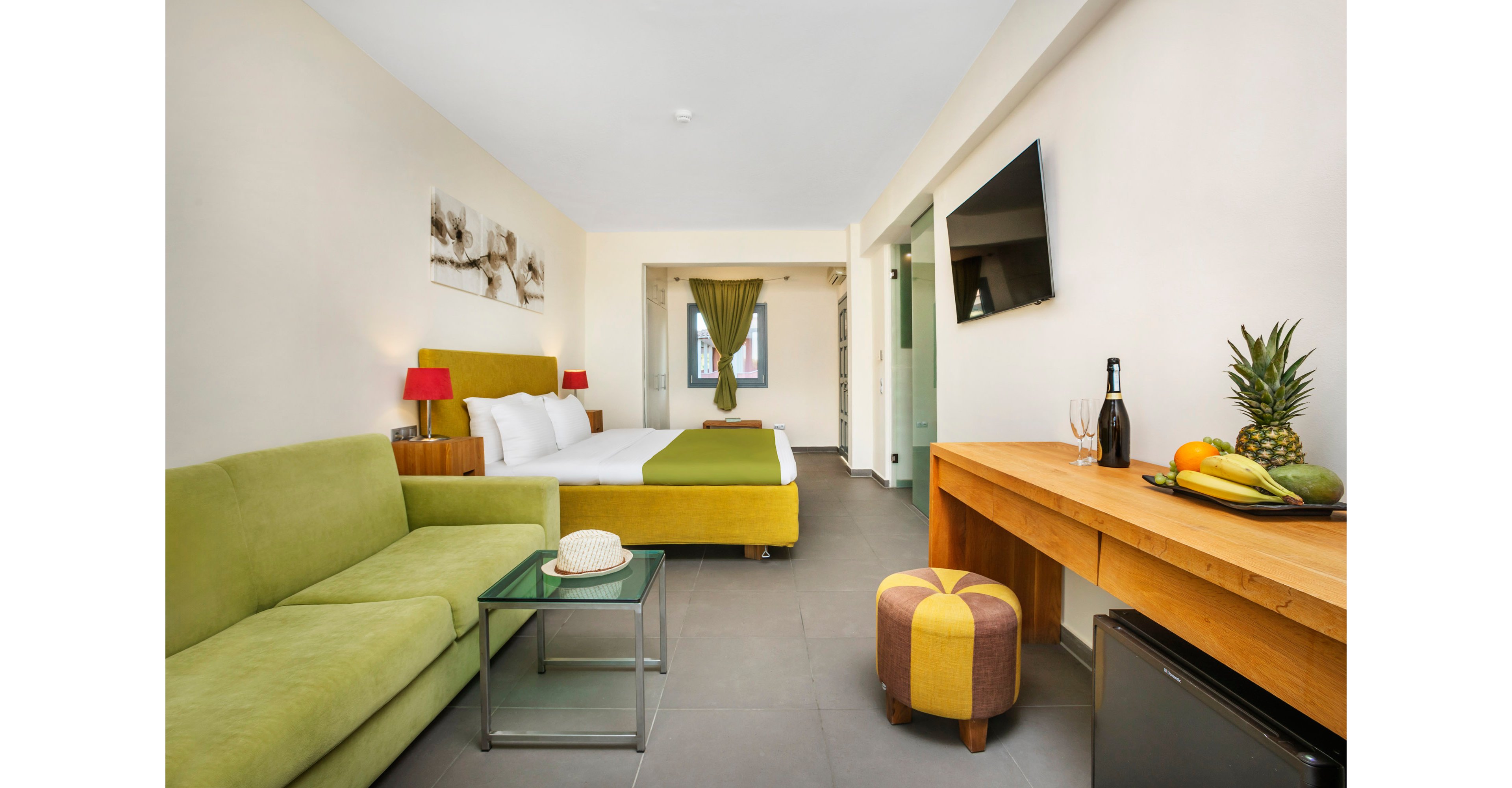 Bright contemporary hotel room featuring a green sofa and matching bed runner. The space includes a double bed with white linens, bedside tables with red lamps, and a wall-mounted flat-screen TV. A wooden desk holds a fruit arrangement, wine bottle, and glasses, while a small ottoman and glass coffee table complete the seating area. Neutral walls and tiled flooring create a clean, minimalist atmosphere with colorful decor highlights.