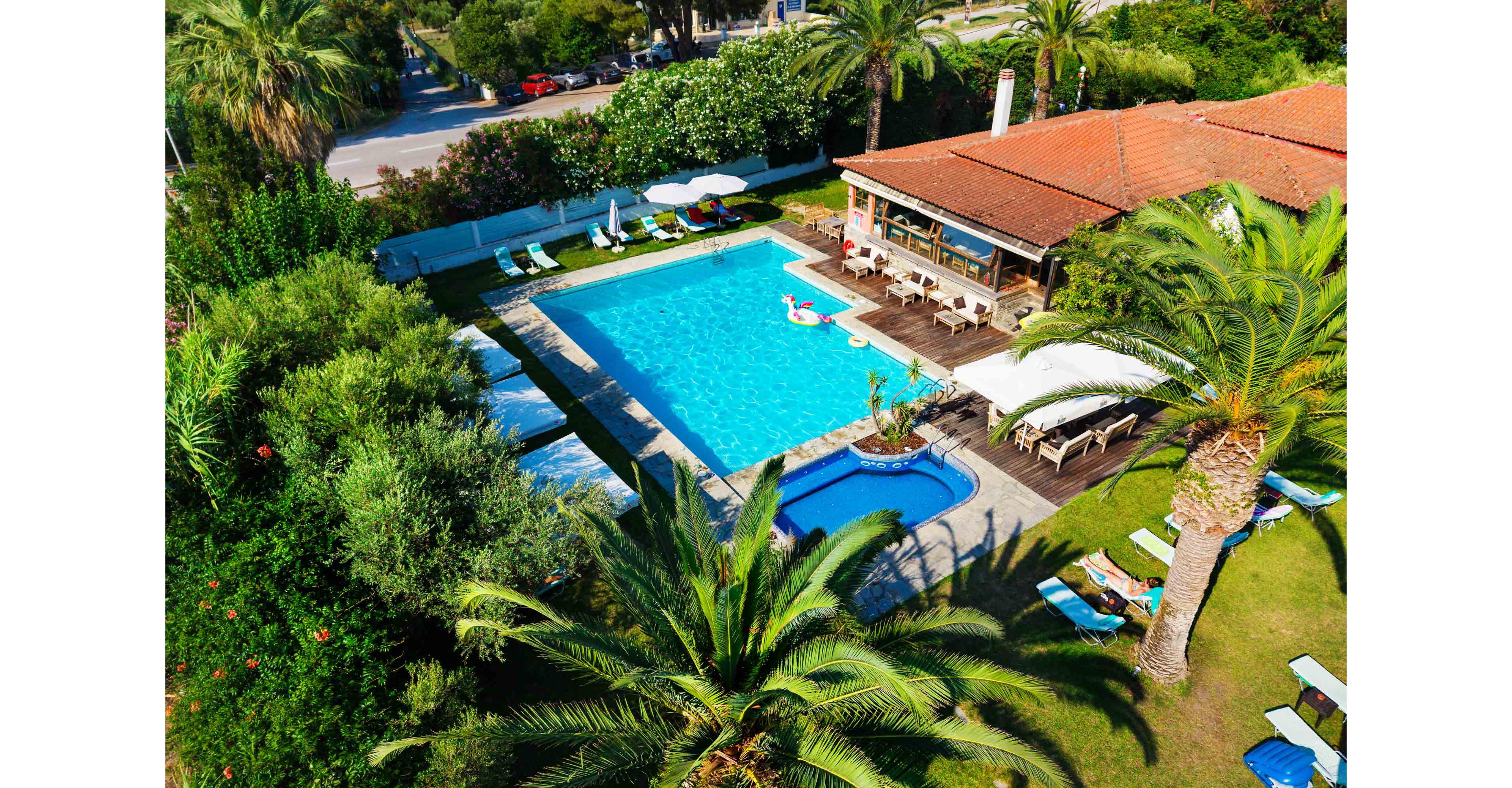 A bright blue swimming pool is the centerpiece of a lush tropical villa property seen from above. Sun loungers and umbrellas surround the main pool and a smaller adjacent pool, creating a relaxing resort atmosphere. Palm trees and dense greenery frame the scene, while a red-roofed building with an outdoor terrace sits beside the water. The vivid colors and clear weather suggest a sunny vacation destination ideal for leisure and relaxation.