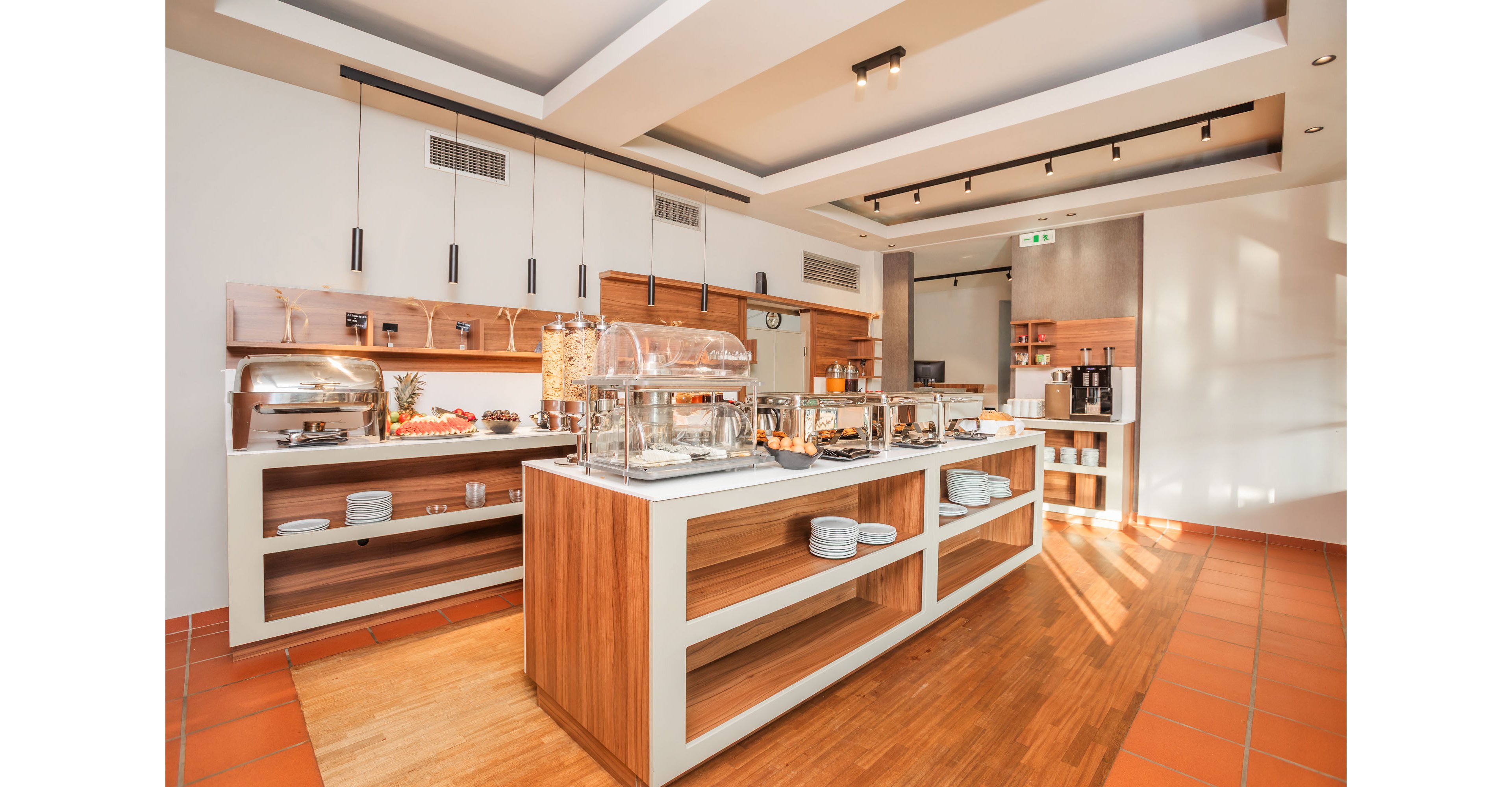 A bright, modern buffet station is set up in a hotel or restaurant dining area. The wooden and white counter displays covered chafing dishes, cereal dispensers, and assorted breakfast items. Clean plates and bowls are neatly stacked on open shelves below the serving surface. Warm lighting, tiled flooring, and minimalist decor create an inviting, contemporary atmosphere.