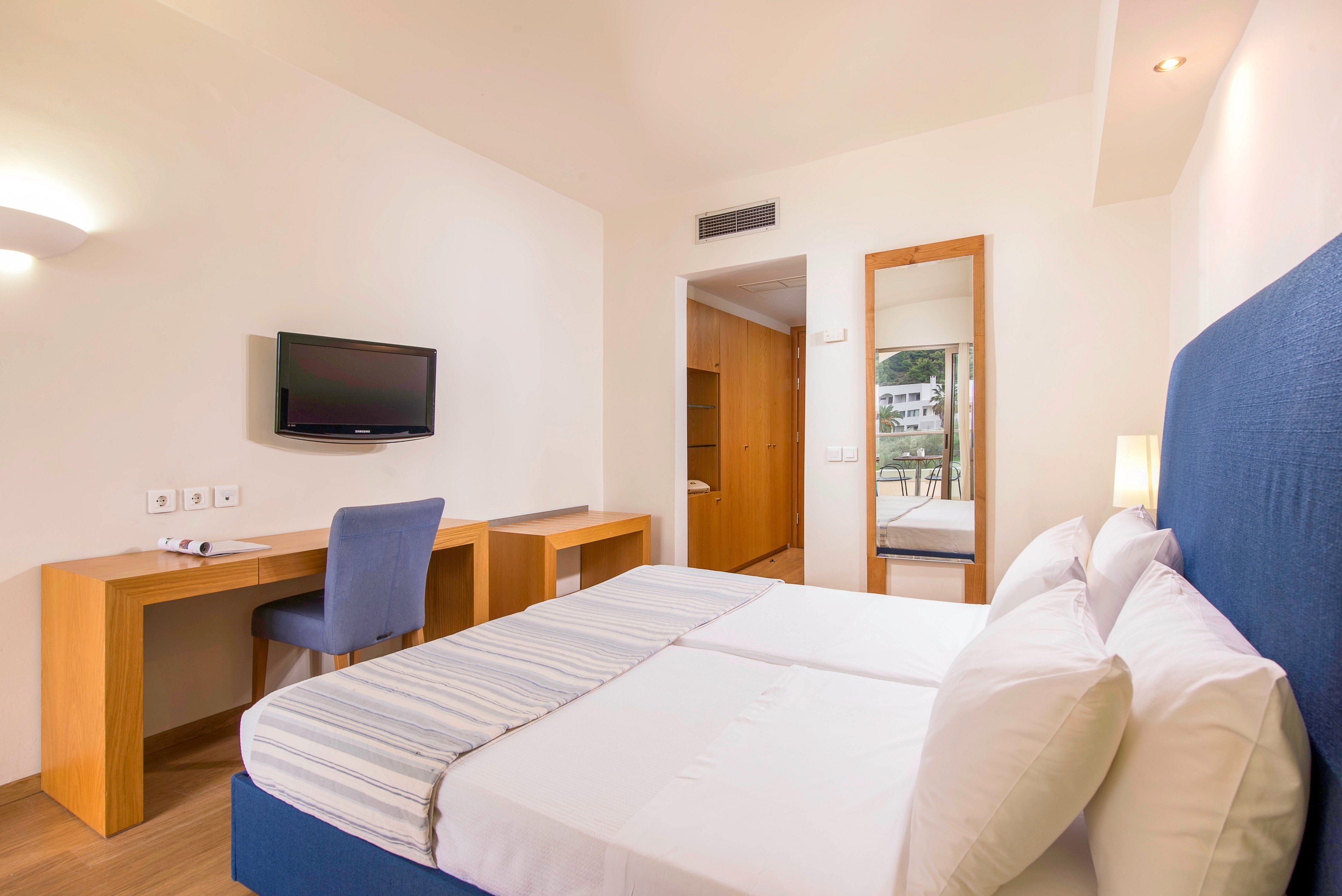 Bright contemporary hotel room featuring a double bed with white linens and a blue upholstered headboard. A wooden desk with a blue chair faces a wall-mounted flat-screen television. A tall mirror and open doorway reveal additional space and natural light from outside. The room is decorated in a minimalist style with neutral walls and soft lighting, creating a clean and inviting atmosphere.