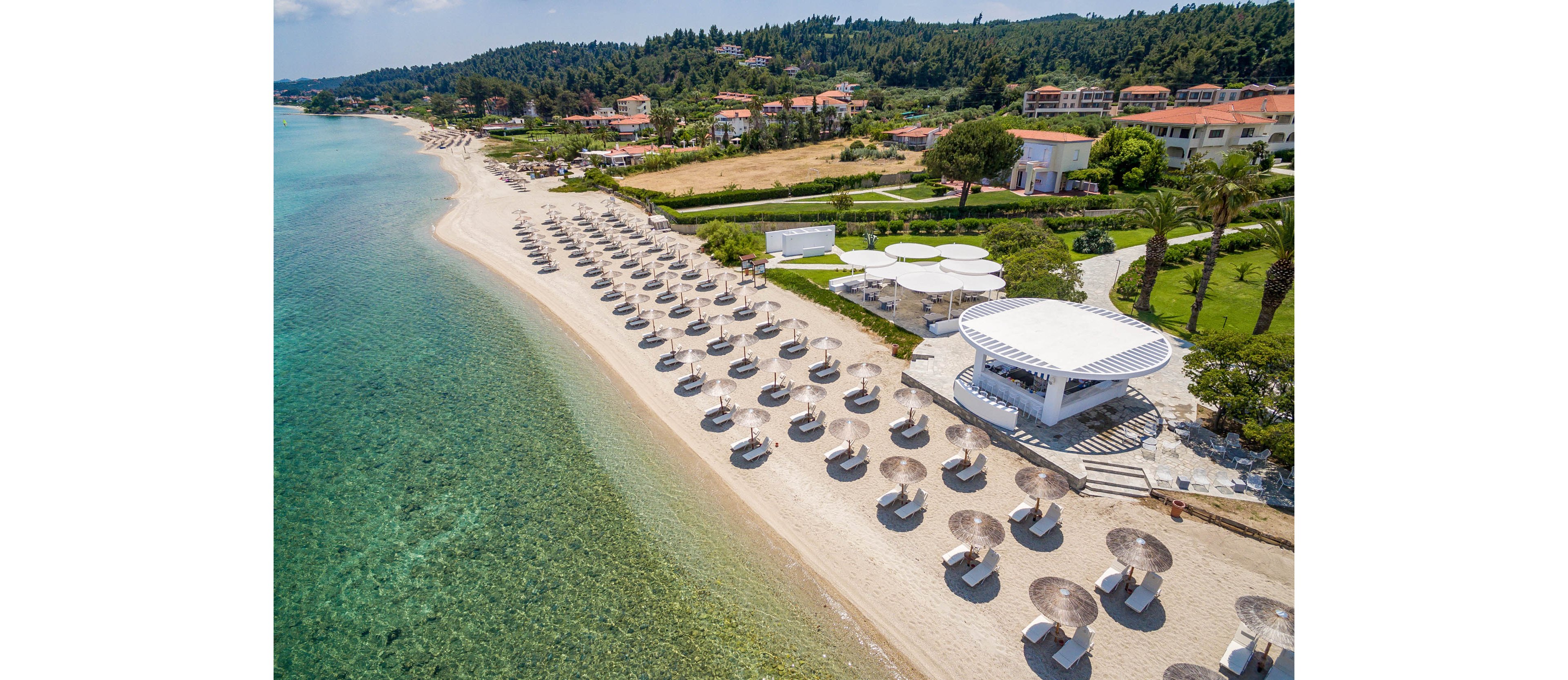 Aerial photograph of a long sandy beach lined with rows of sun loungers and parasols beside clear turquoise sea. A modern white beachfront pavilion sits near the shoreline, surrounded by landscaped gardens and resort buildings. The water transitions from light to deep blue, highlighting the gentle slope of the coast. In the background, green hills and villas create a relaxed holiday atmosphere.