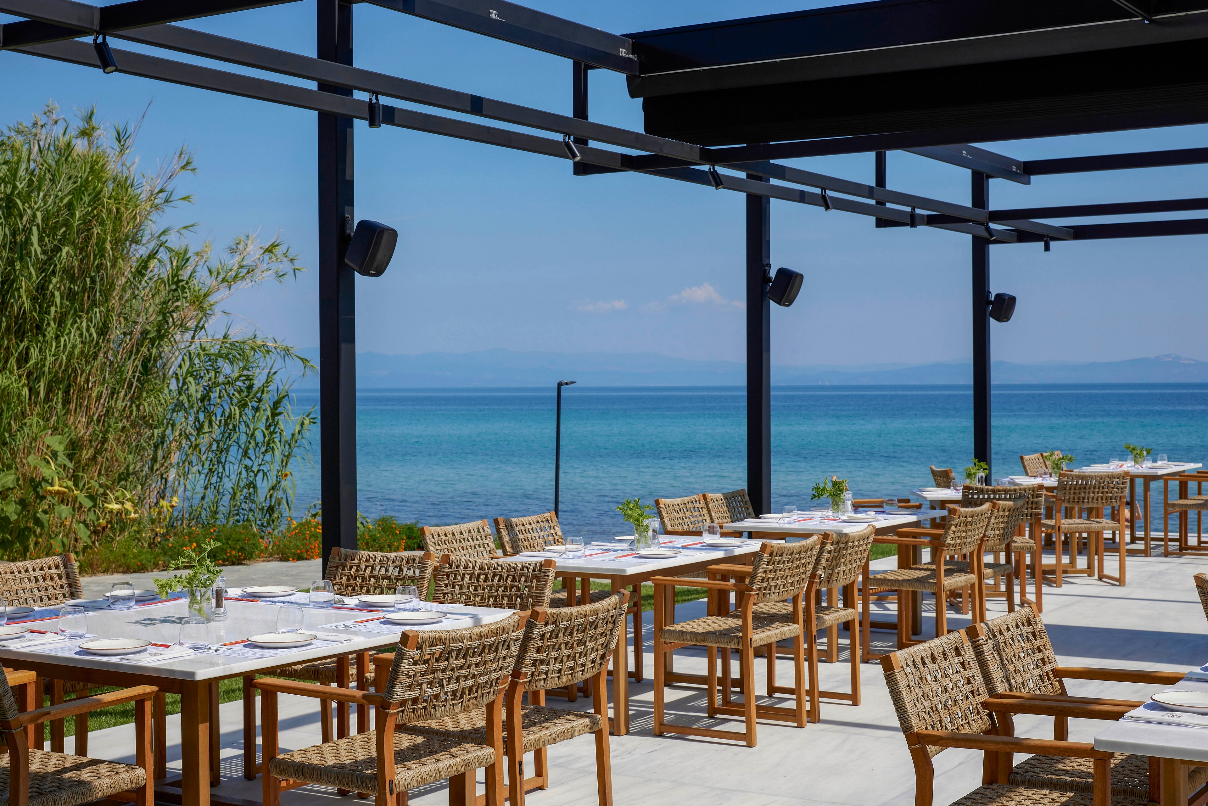 An open-air restaurant terrace overlooks a calm blue sea under a clear sky. Wooden tables and woven chairs are neatly arranged in rows, set with plates and glassware for guests. A modern pergola structure frames the seating area, casting geometric shadows on the light stone floor. Lush green plants border the terrace, adding a natural touch to the coastal setting.