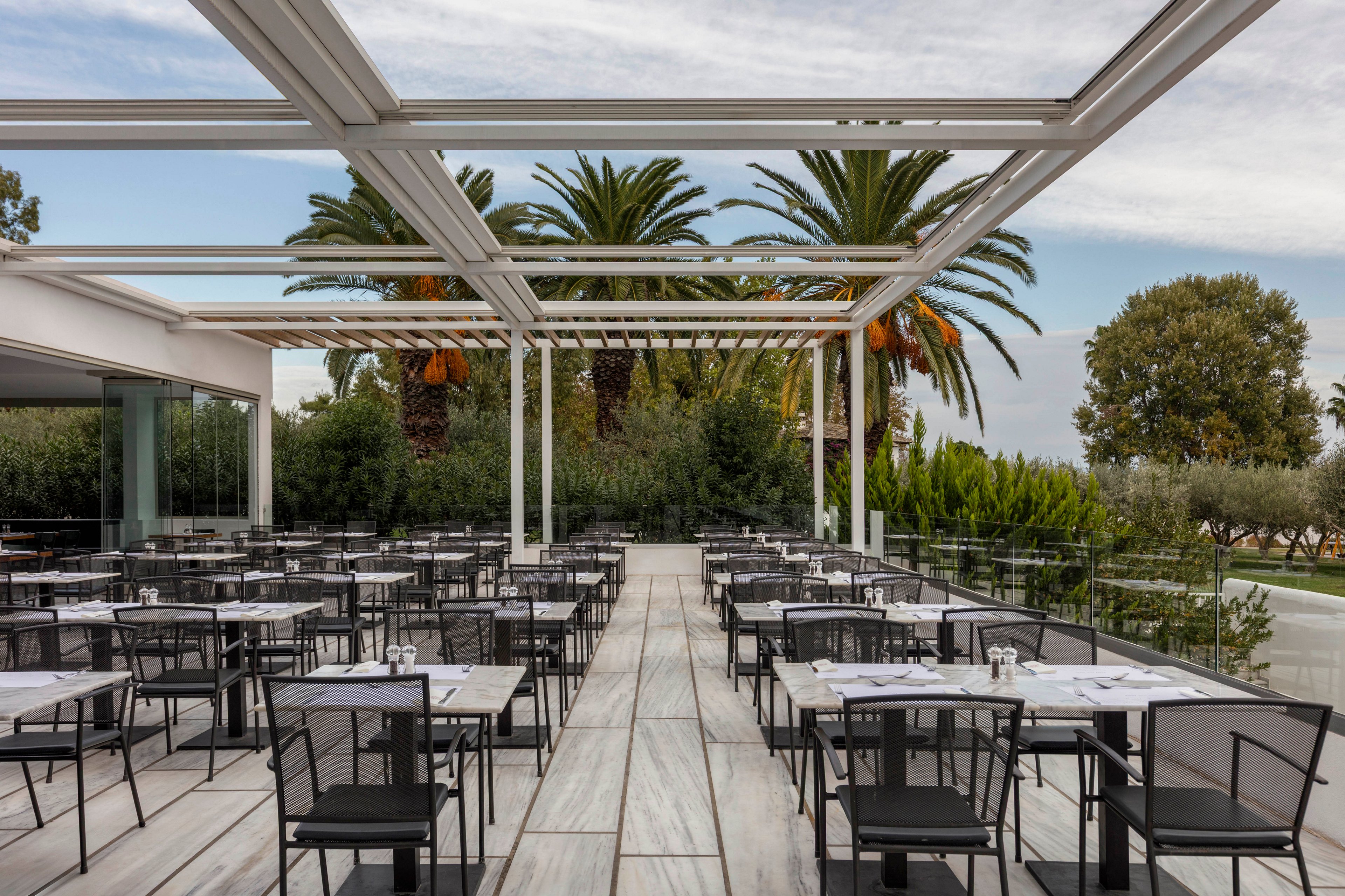 A contemporary open-air restaurant terrace is arranged with rows of black metal tables and chairs, each set with white tablecloths and glassware. The space features a sleek pergola structure above light stone flooring, creating a clean, minimalist look. Lush greenery and tall palm trees frame the background, suggesting a warm, resort-like environment. The scene is calm and orderly, with no people present, emphasizing the architectural design and outdoor dining atmosphere.