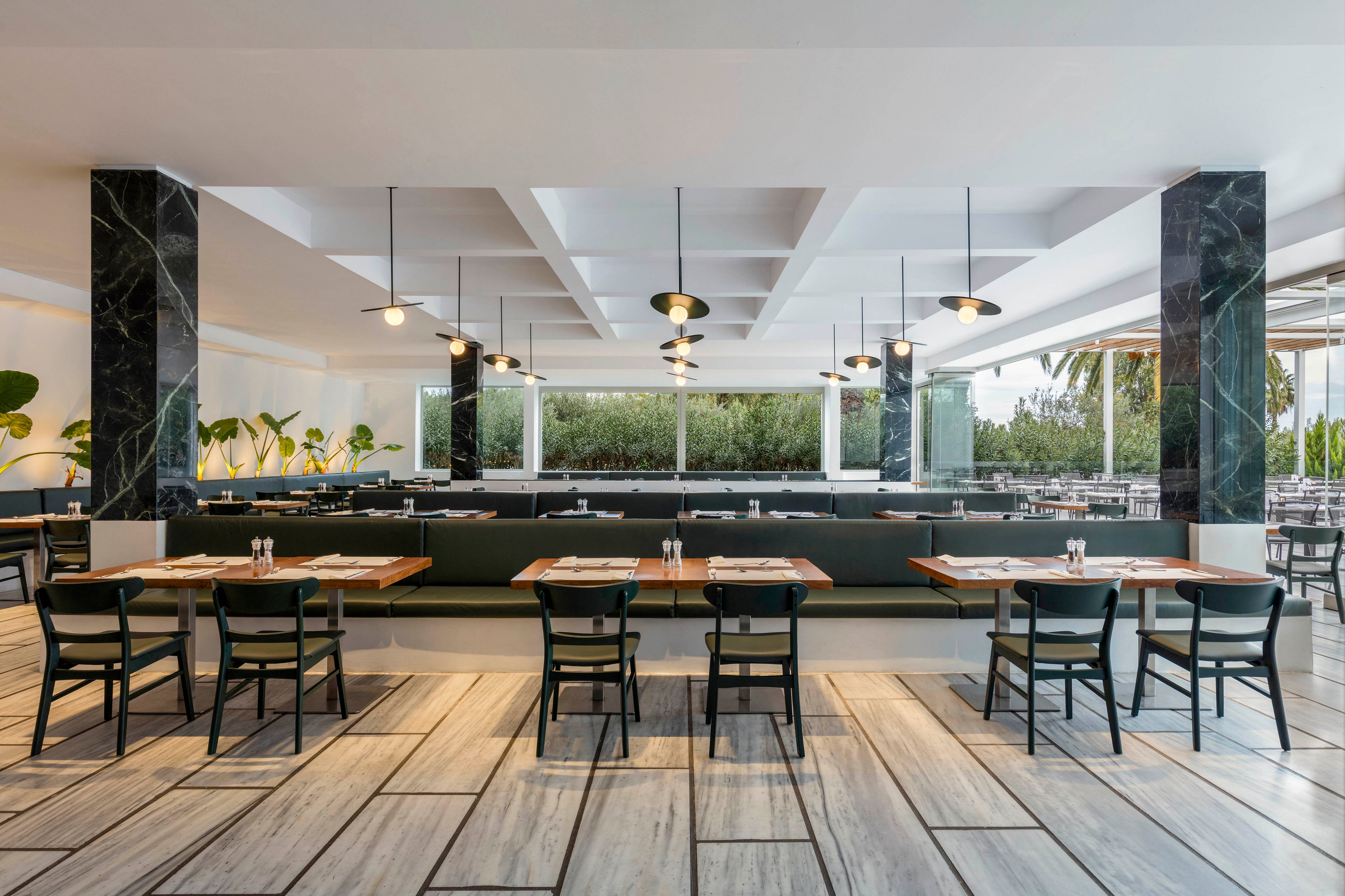 Contemporary restaurant dining area with neatly arranged wooden tables and dark chairs is shown empty and ready for guests. The space features a long central banquette, pendant lights, and large windows opening to an outdoor seating area. Light-colored tiled flooring with dark lines adds a geometric pattern, while plants and greenery outside bring a natural touch. Overall, the scene conveys a clean, minimalist, and inviting atmosphere.
