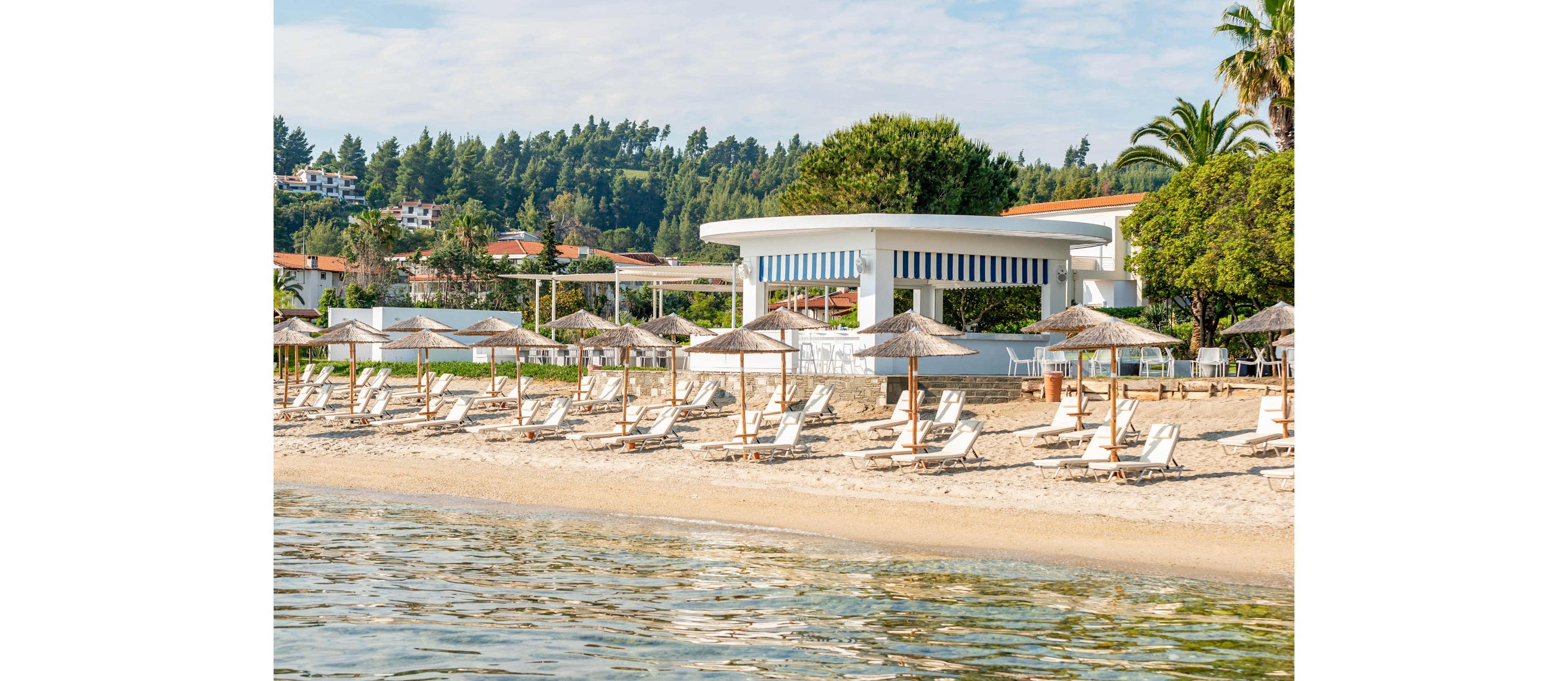 A tranquil sandy beach is lined with wooden sun loungers and straw parasols facing calm, clear water. In the background, a modern white beachfront pavilion stands among palm trees and lush greenery. Resort buildings are visible on the hillside, suggesting a coastal holiday destination. The overall scene feels relaxed and inviting, ideal for summer vacations and seaside leisure.