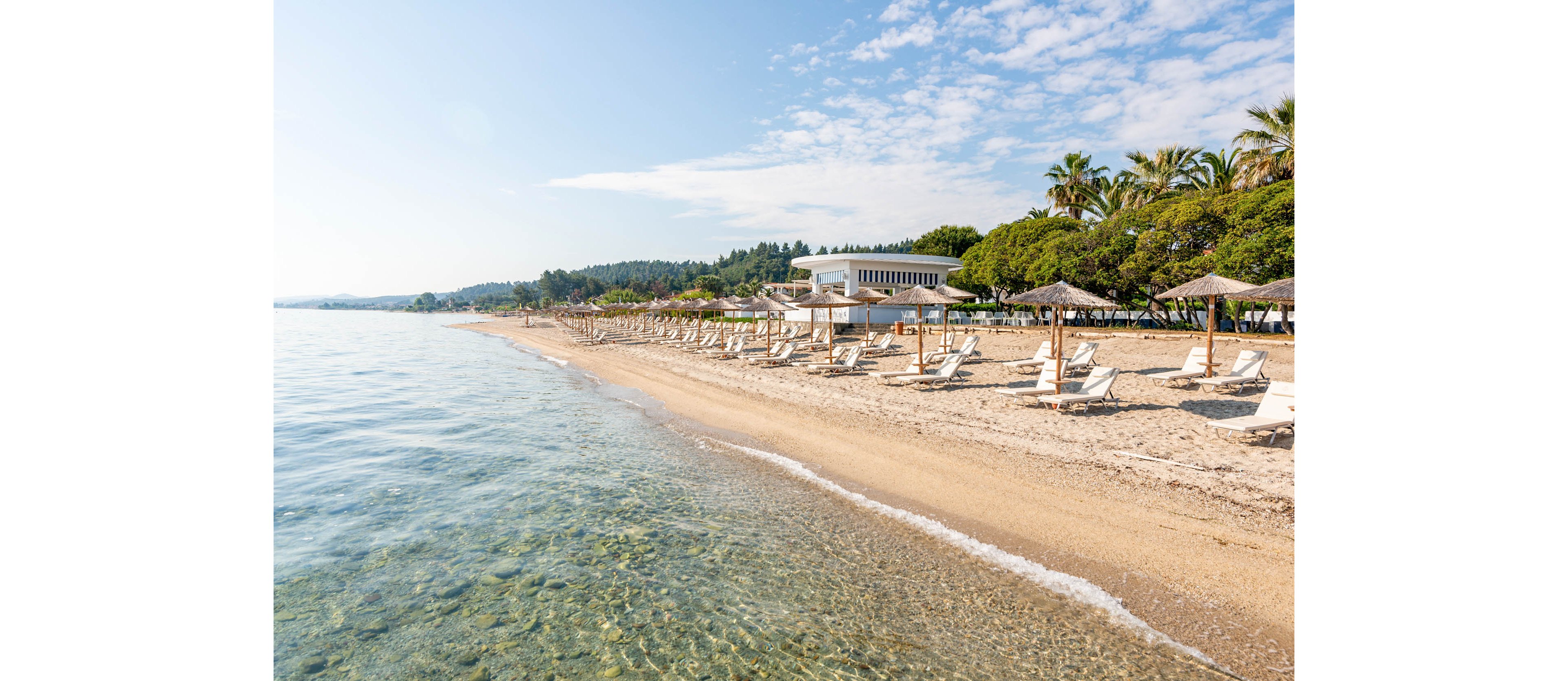 A tranquil sandy beach stretches along clear, shallow coastal waters under a bright blue sky. Rows of sun loungers and straw umbrellas line the shore, suggesting a well-equipped seaside resort. A modern white building is nestled among lush green trees and palm trees in the background. The calm sea, gentle waves, and warm light create a relaxed, inviting holiday atmosphere.