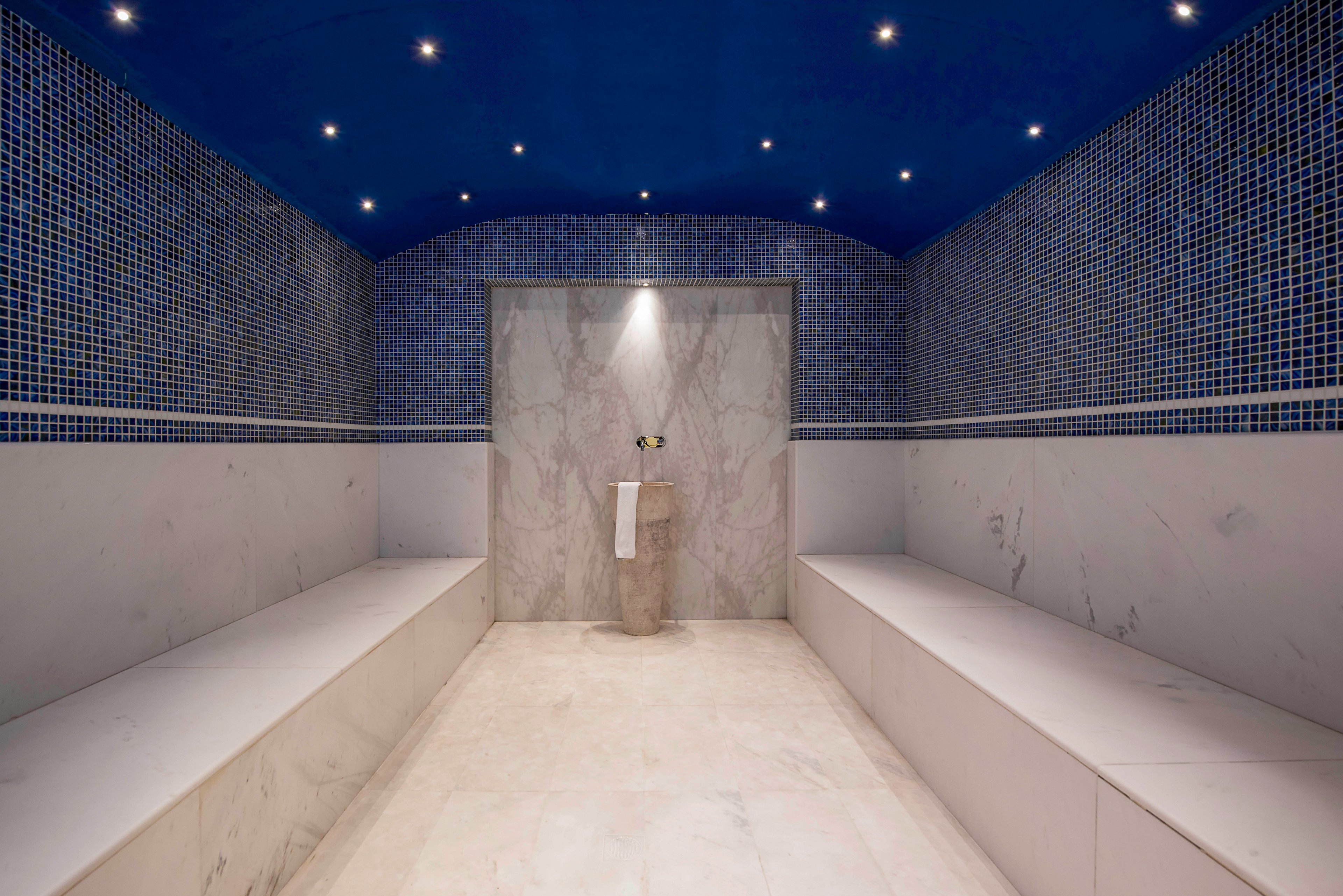 A contemporary steam room features long marble benches along both sides and a smooth marble floor. The back wall has a central pedestal with a folded white towel and a small bowl beneath a spotlight. Blue mosaic tiles cover the upper walls and arched ceiling, accented by multiple recessed lights that create a calm, spa-like atmosphere. The space appears clean, minimal, and designed for relaxation and wellness.