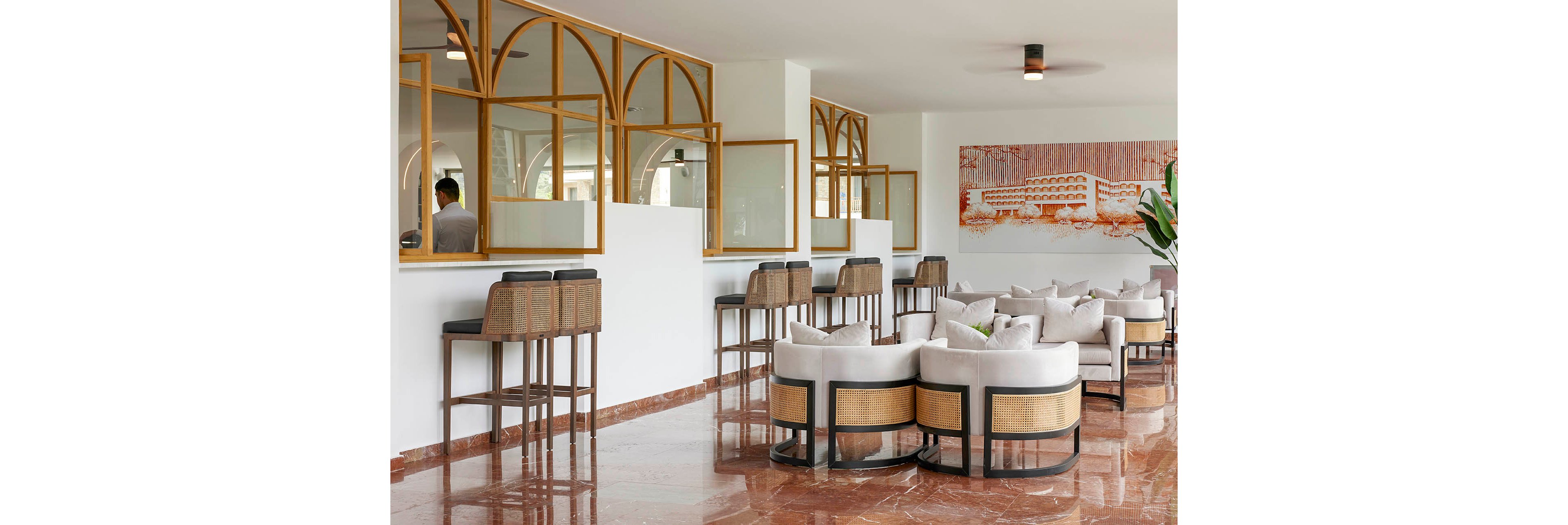 Contemporary hotel lobby interior featuring circular lounge chairs with light upholstery and woven panels. High bar stools line a counter with arched wooden-framed windows, where a staff member is partially visible. The space has glossy brown marble flooring, white walls, and minimalist ceiling lights. A large wall artwork and indoor plant add decorative accents to the clean, modern design.
