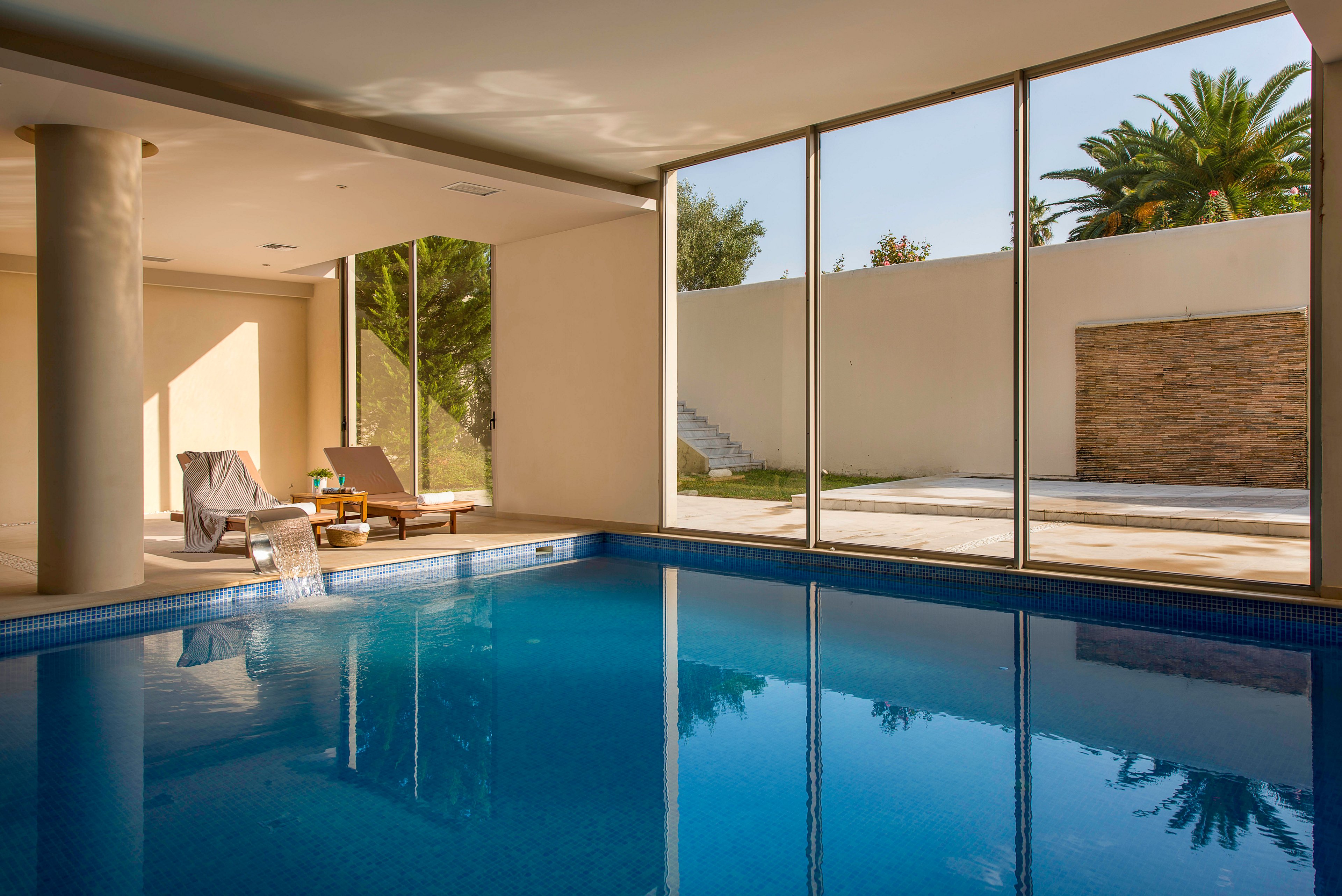 A serene indoor swimming pool reflects the soft natural light streaming through large floor-to-ceiling windows. Two lounge chairs with towels and a small side table are arranged beside the pool, suggesting a relaxing leisure setting. Outside, a minimalist patio, stone wall, and lush palm trees create a modern, resort-like atmosphere. The overall scene feels calm and inviting, emphasizing comfort and tranquility.