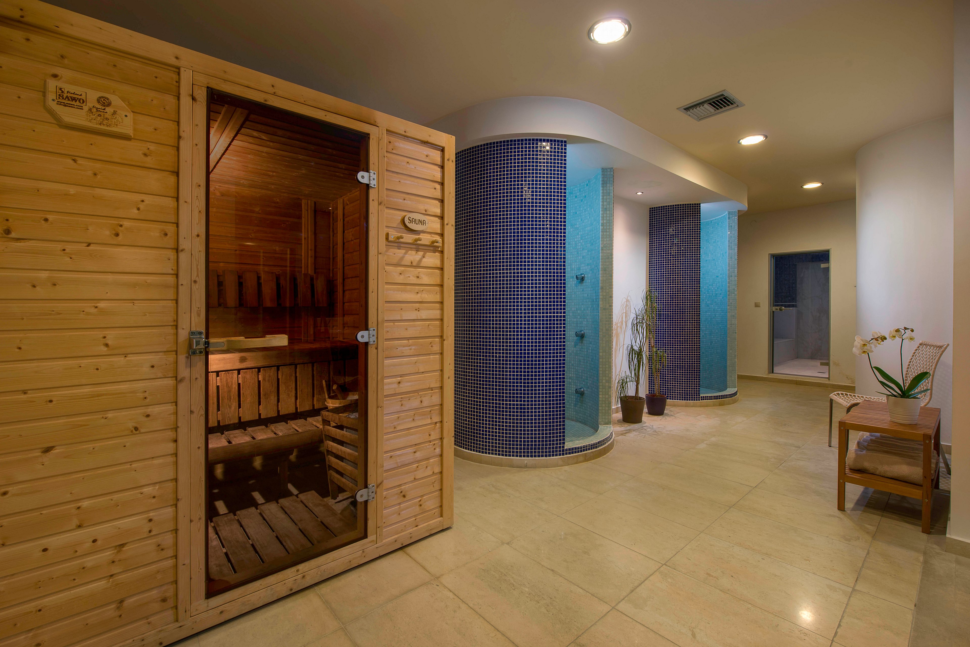 A contemporary spa interior features a wooden sauna cabin on the left side of the room. In the center, a curved column covered with small blue tiles adds a decorative accent near a plant and additional spa facilities. The space has smooth beige flooring, soft ambient lighting, and neutral walls that create a calm atmosphere. A simple wooden chair with a flower arrangement sits along the right wall, enhancing the relaxing, minimalist design.