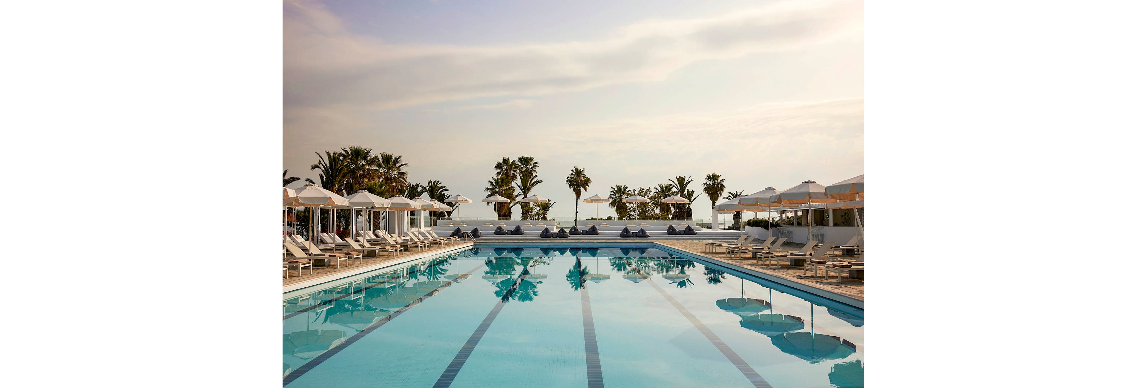 A large outdoor swimming pool is surrounded by white lounge chairs and umbrellas at a luxury seaside resort. Palm trees line the far edge of the pool, framing a view of the ocean in the background. The calm water reflects the soft evening sky, creating a tranquil and inviting atmosphere. The scene suggests relaxation and vacation leisure in a warm coastal destination.