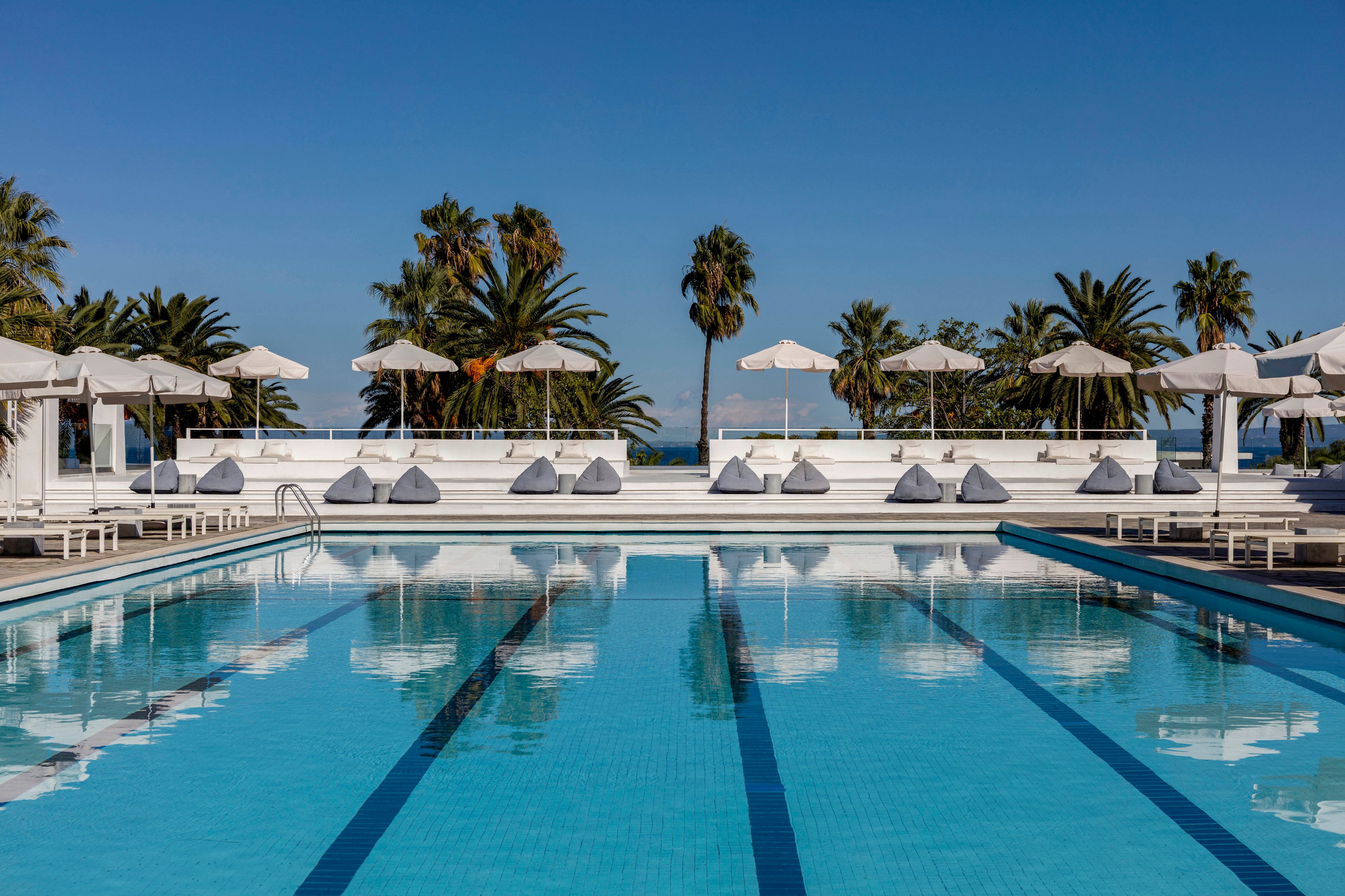 A large, clear blue swimming pool stretches toward a row of white sun loungers and umbrellas. Cushioned daybeds line the far side of the pool, creating a relaxed resort atmosphere. Tall palm trees rise behind the seating area under a bright, cloudless sky. The scene suggests a tranquil, upscale outdoor leisure setting near the coast.