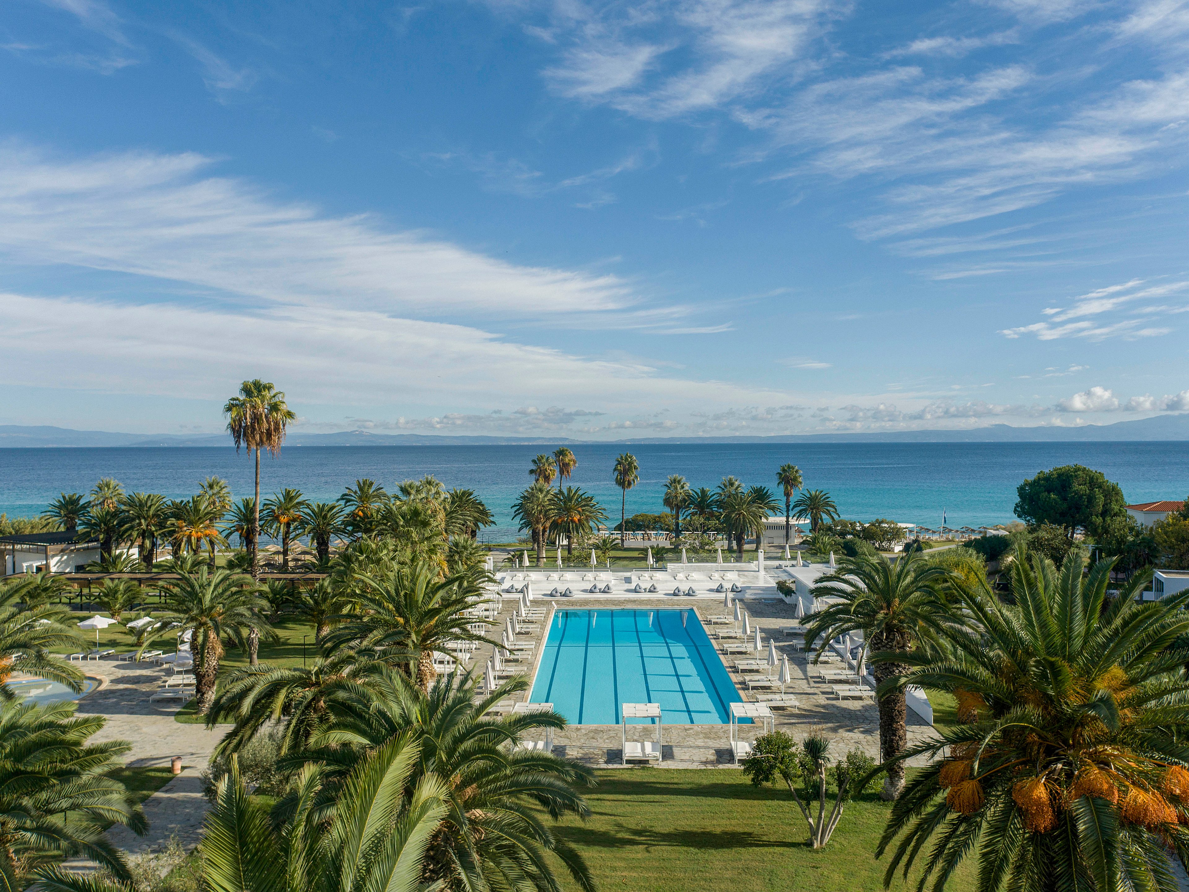 A large rectangular swimming pool is centered in a tropical resort overlooking a calm blue sea. Palm trees and manicured lawns surround the pool area, with sun loungers neatly arranged along the deck. The horizon shows distant landforms under a bright sky with scattered clouds. The scene conveys a tranquil, sunny vacation atmosphere by the coast.