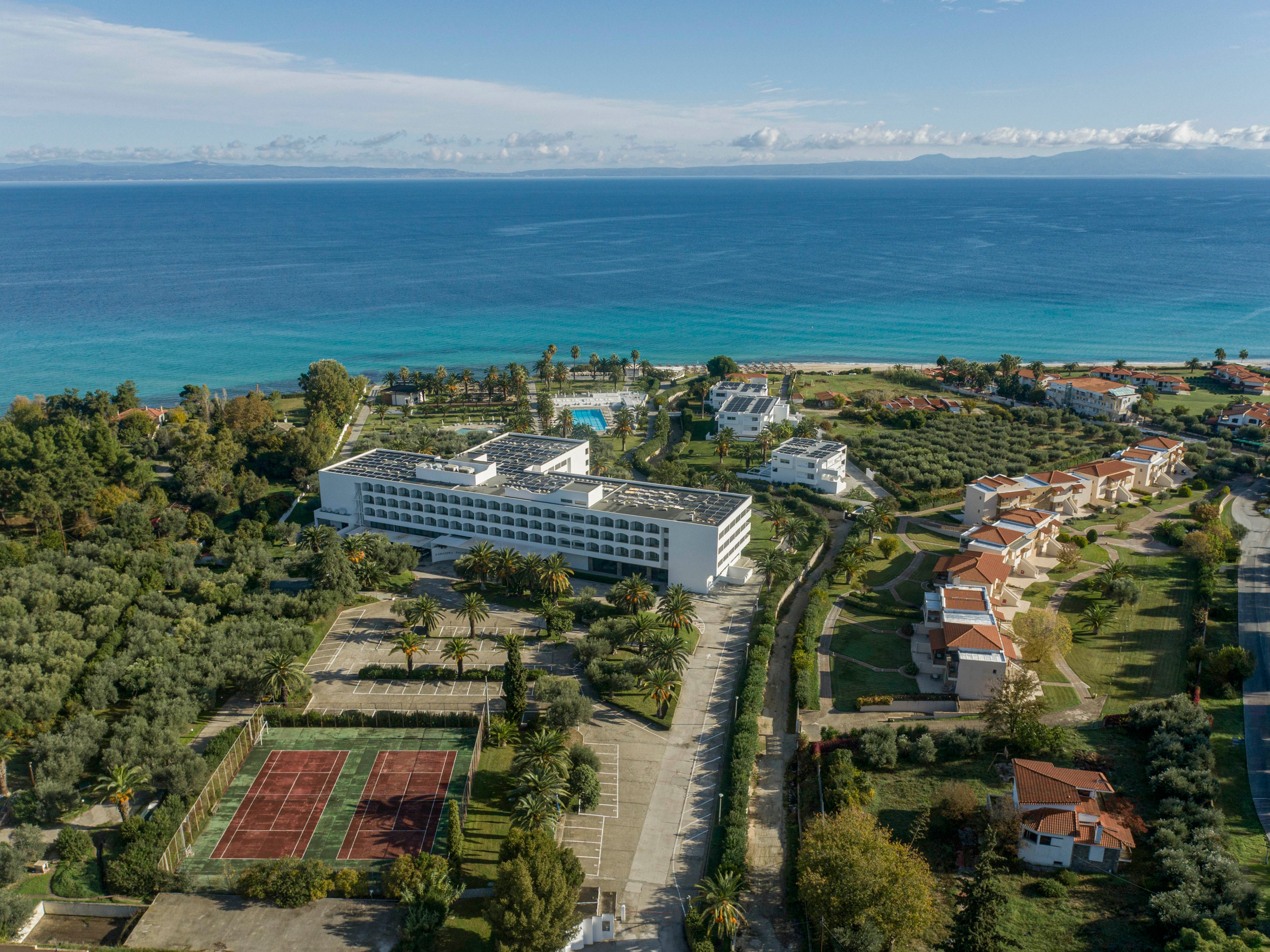 A large modern resort complex is situated along a bright blue coastline, surrounded by lush greenery and trees. Multiple white buildings and villas are spread across the property, with red tennis courts visible in the foreground. The scene is captured from an elevated viewpoint, showing the expansive grounds and the calm sea stretching to the horizon. Clear skies and soft clouds contribute to a serene, vacation-like atmosphere.