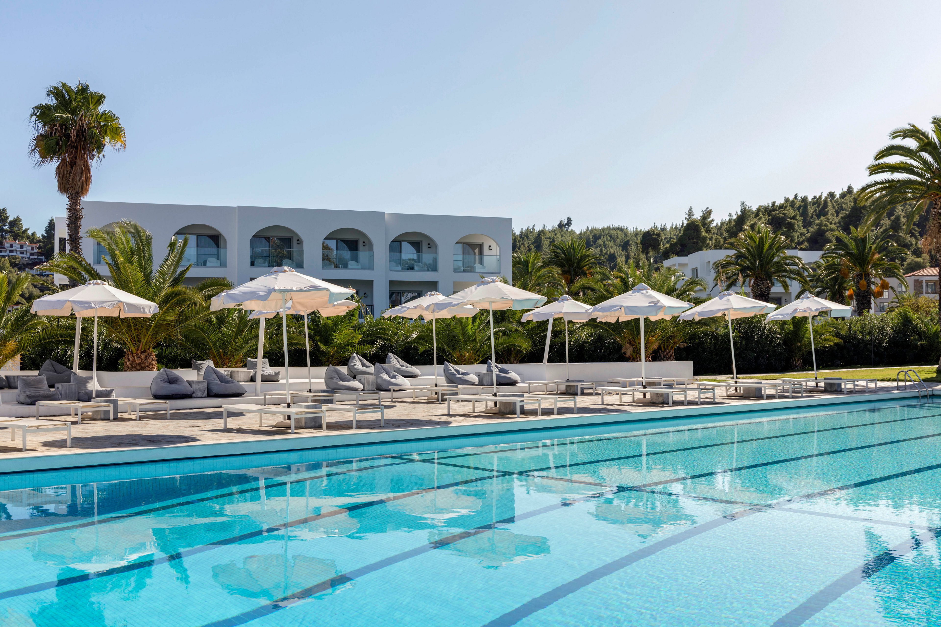 A large outdoor swimming pool reflects the clear blue sky at a modern resort. White sun umbrellas and cushioned loungers line the poolside, creating a relaxed vacation atmosphere. In the background, a white building with arched balconies is surrounded by lush green palm trees and vegetation. The scene suggests a tranquil, sunny day ideal for leisure and relaxation.