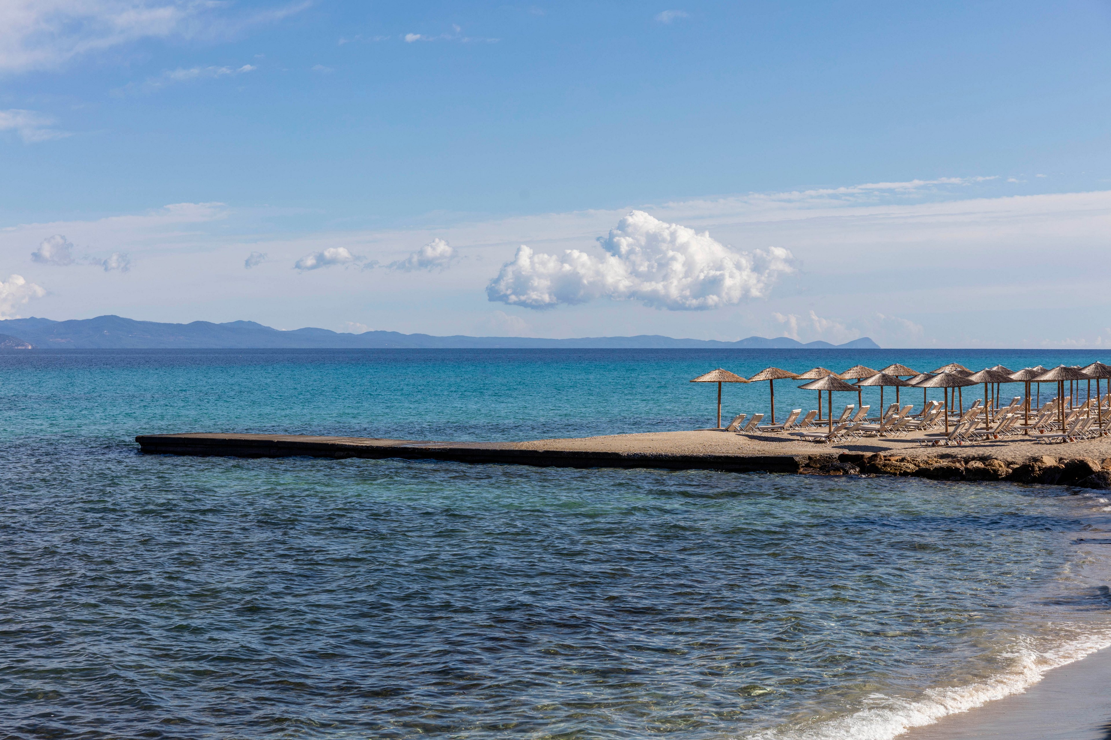 A tranquil coastal scene shows a small pier extending into clear blue sea water. Straw sun umbrellas line the pier, suggesting a quiet beach resort with no people present. Distant mountains are visible on the horizon under a bright blue sky with scattered clouds. Gentle waves lap against the shore, creating a peaceful vacation atmosphere.